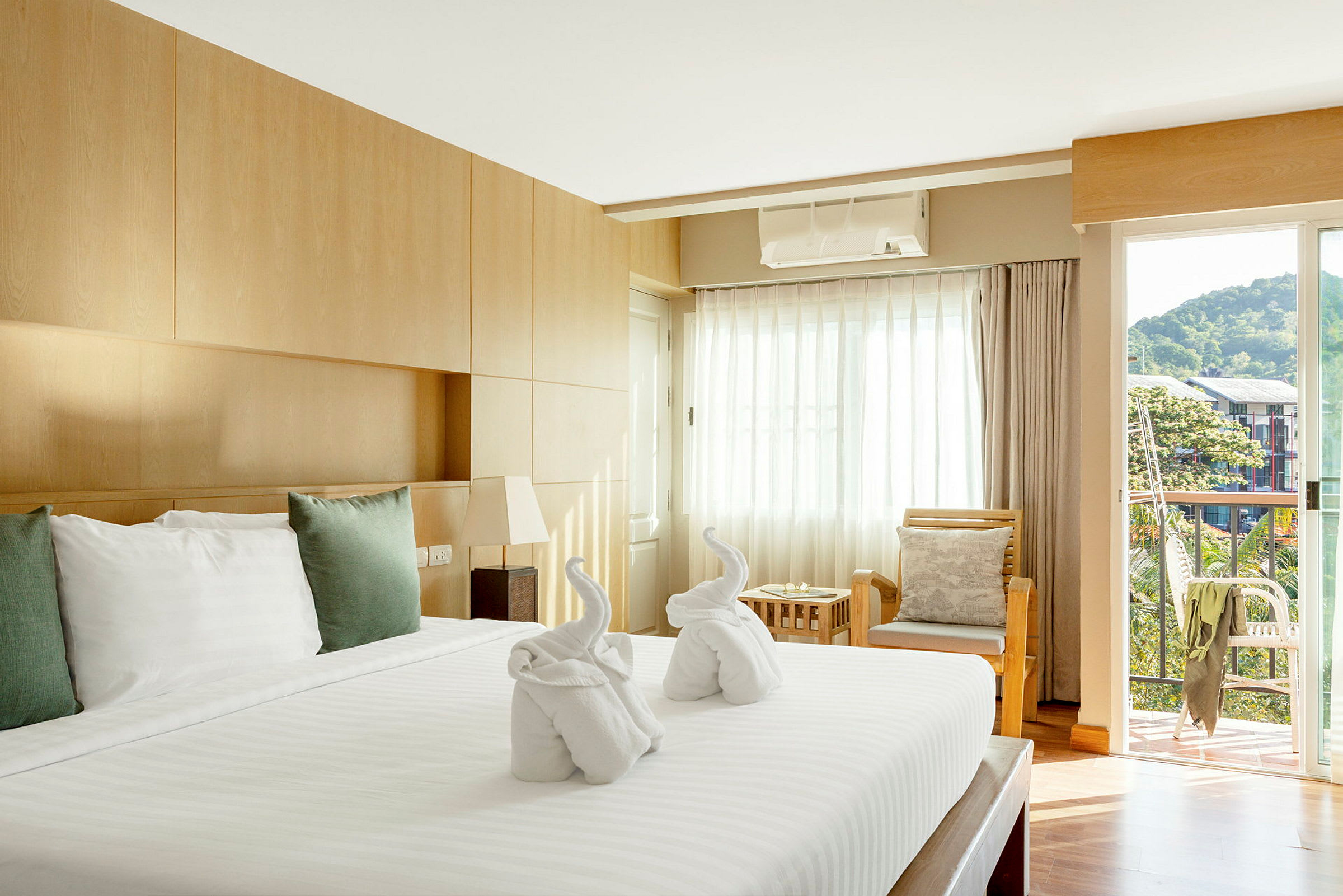 Valoisa hotellihuone, jossa on suuri sänky peitettynä valkoisilla lakanoilla ja tyynyillä. Sängyllä on kaksi pyyhettä taitettuna joutsenten muotoon. Huoneessa on puukalusteita, istuinryhmä tyynyineen ja parveke näkymällä vihreään ympäristöön ja rakennuksiin. (Tekoälyn luoma teksti)