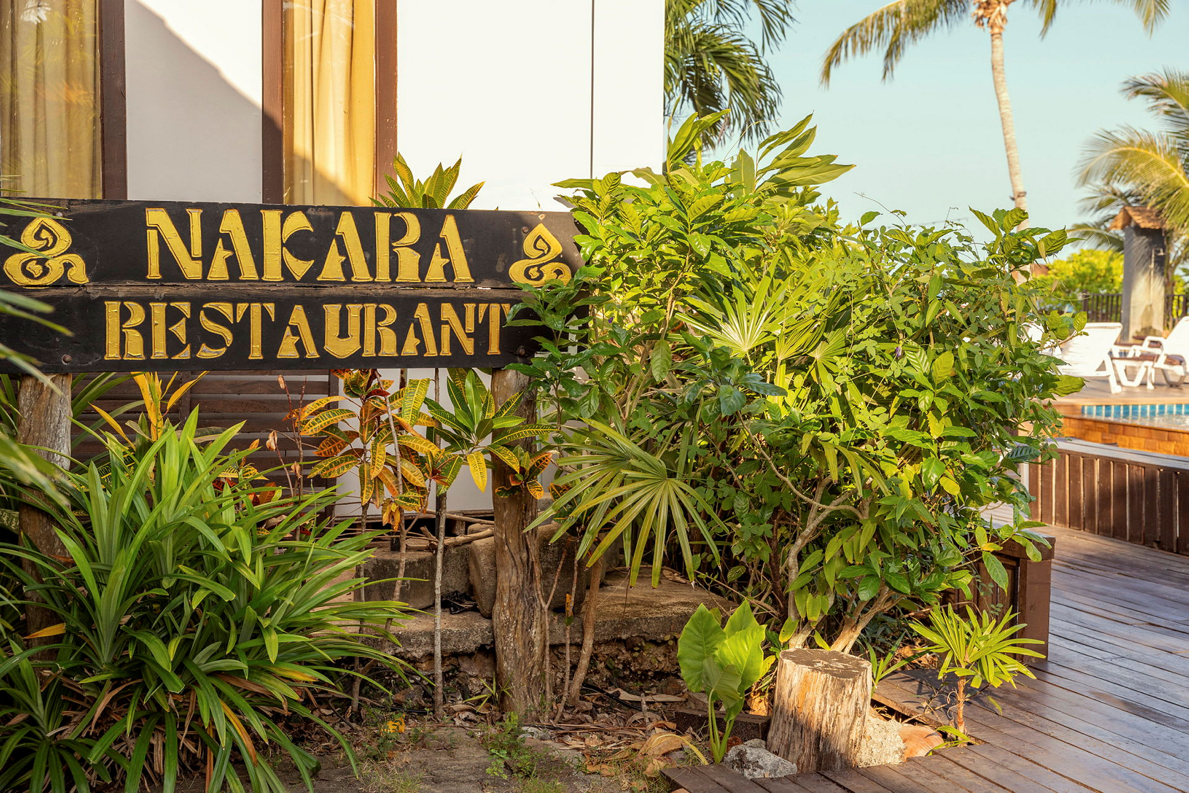 Kyltti, jossa lukee "Nakara Restaurant", ympäröi kasvillisuus, ja taustalla näkyy puualusta ja uima-allas. (Tekoälyn luoma teksti)