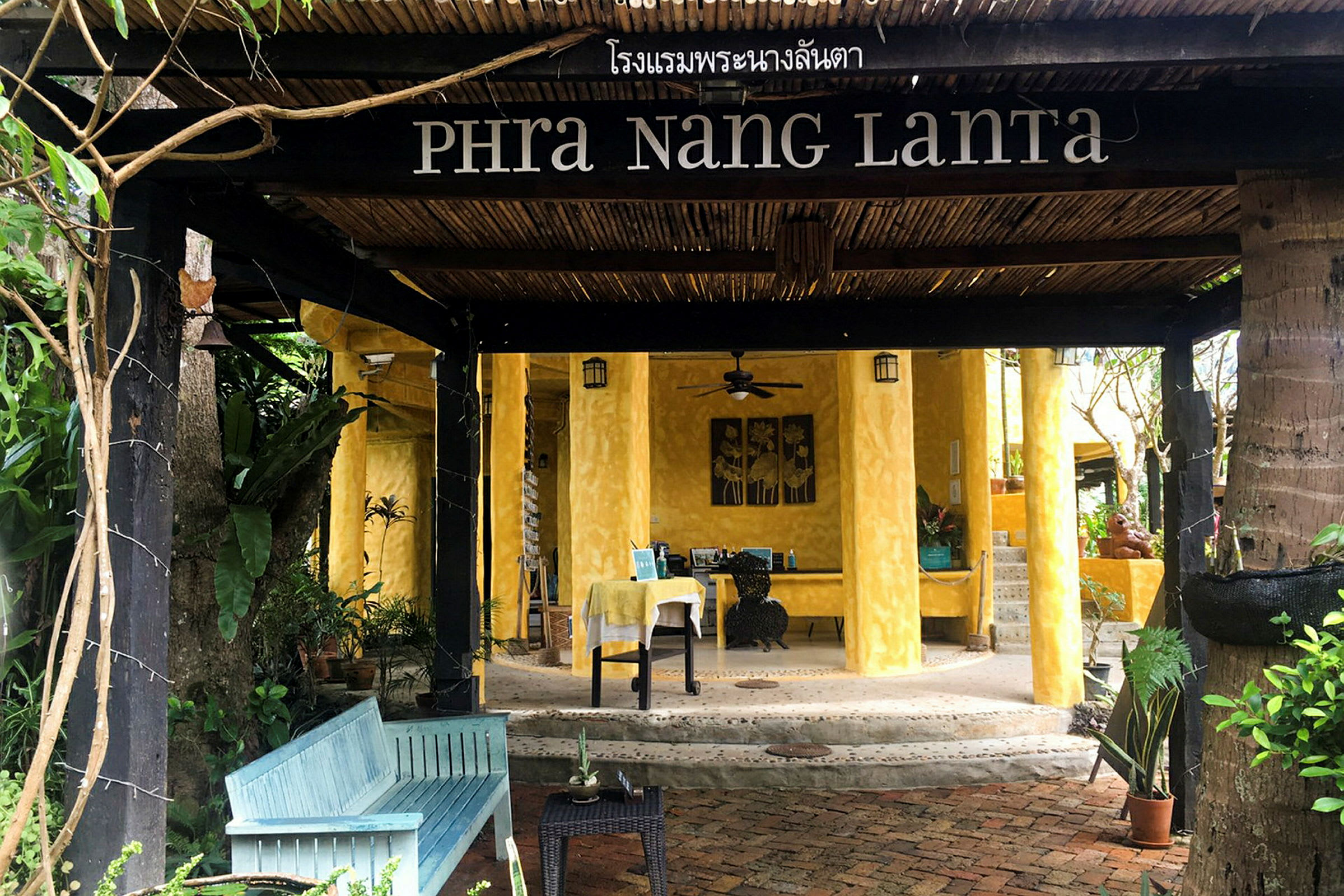 Hotellin sisäänkäynti, jonka nimi on Phra Nang Lanta, keltaisia seiniä ja puuyksityiskohtia, vehreyden ympäröimänä. (Tekoälyn luoma teksti)