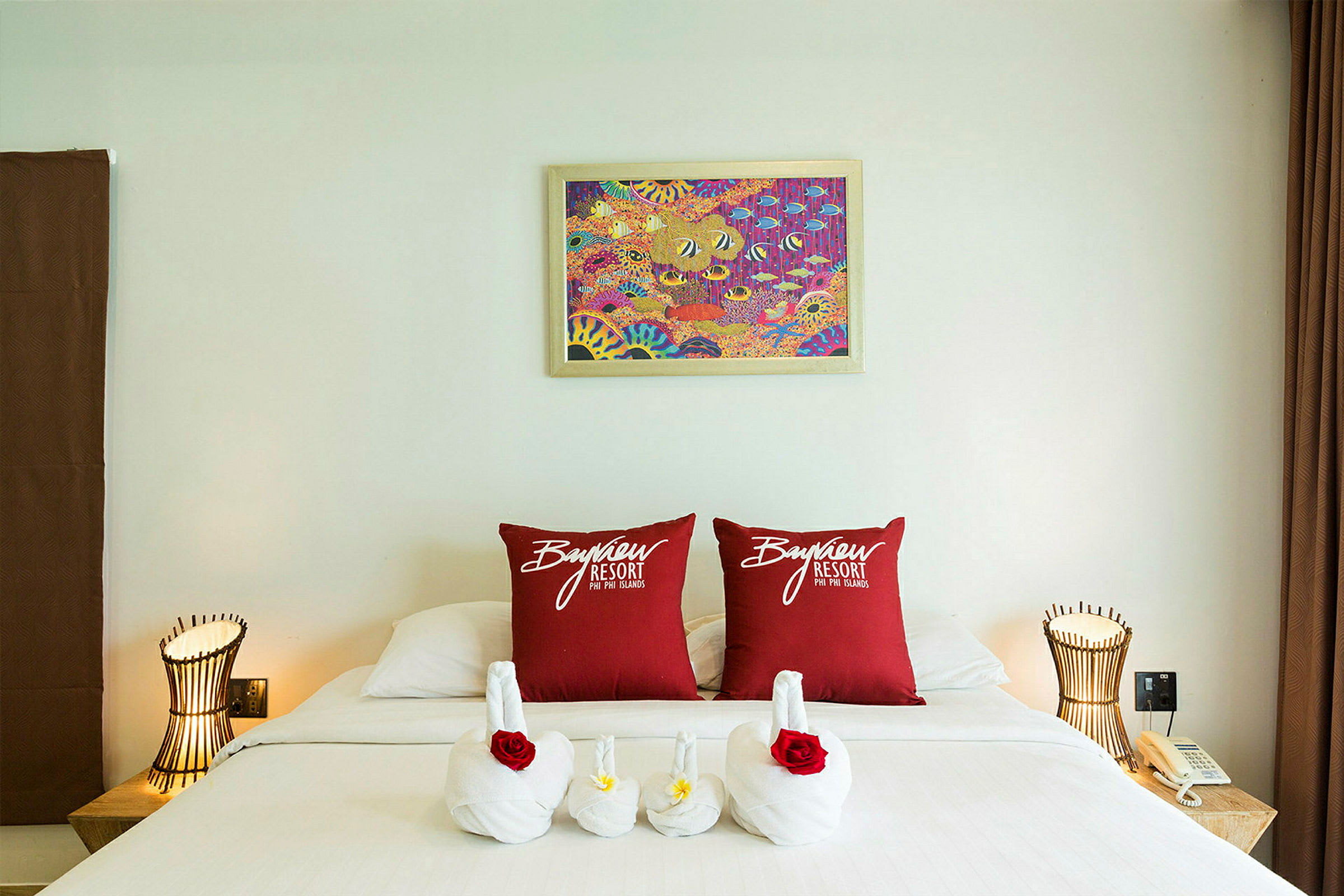 Hotellihuone, jossa on koristeltu sänky. Kaksi punaista tyynyä, joissa lukee "Bayview Resort", on asetettu sängylle, ja pyyhkeet on taiteltu joutseniksi kukilla. Yöpöydällä on lamppu ja puhelin. (Tekoälyn luoma teksti)