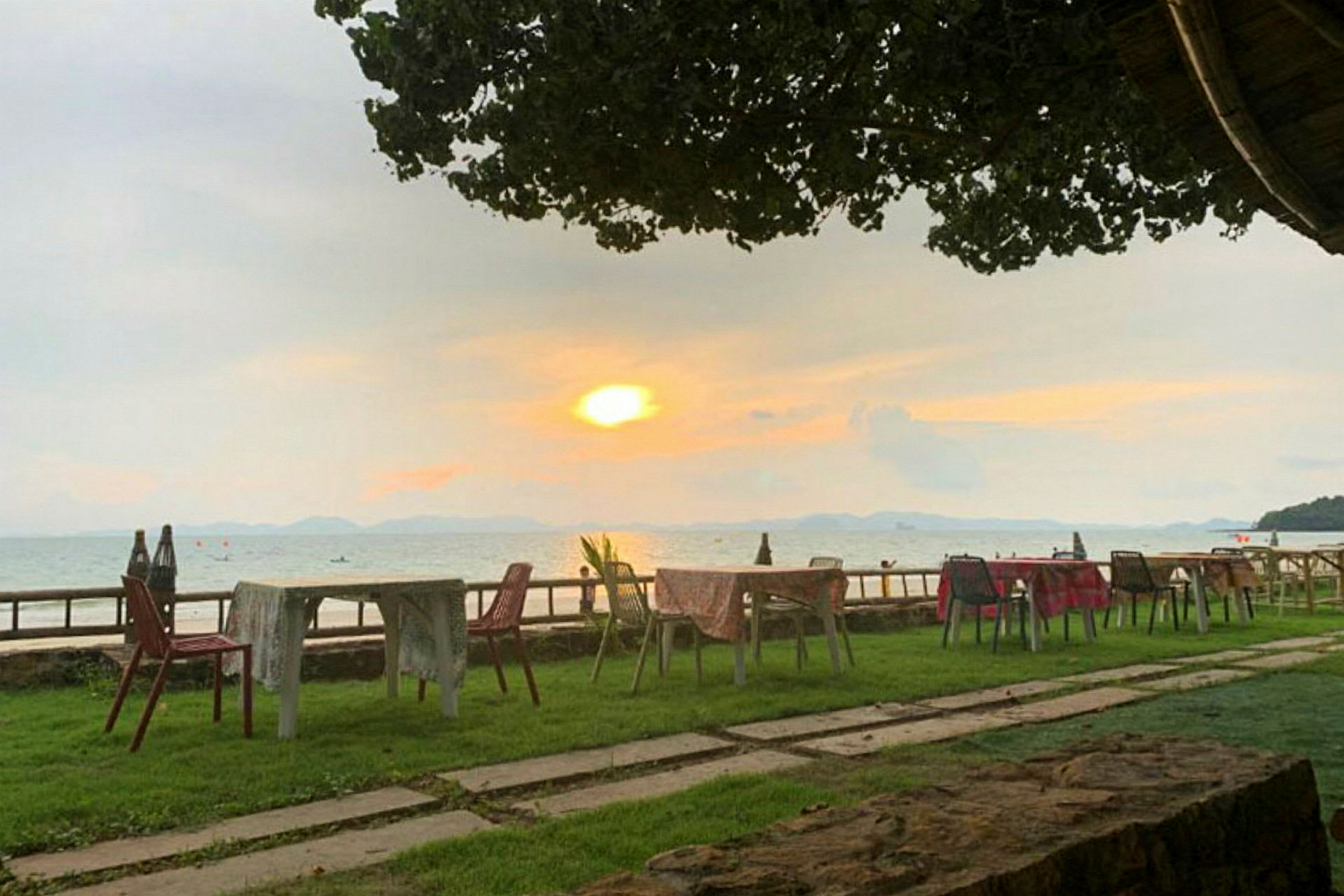 Ulkoterassi, jossa pöytiä ja tuoleja nurmikolla meren äärellä ja taustalla auringonlasku. (Tekoälyn luoma teksti)