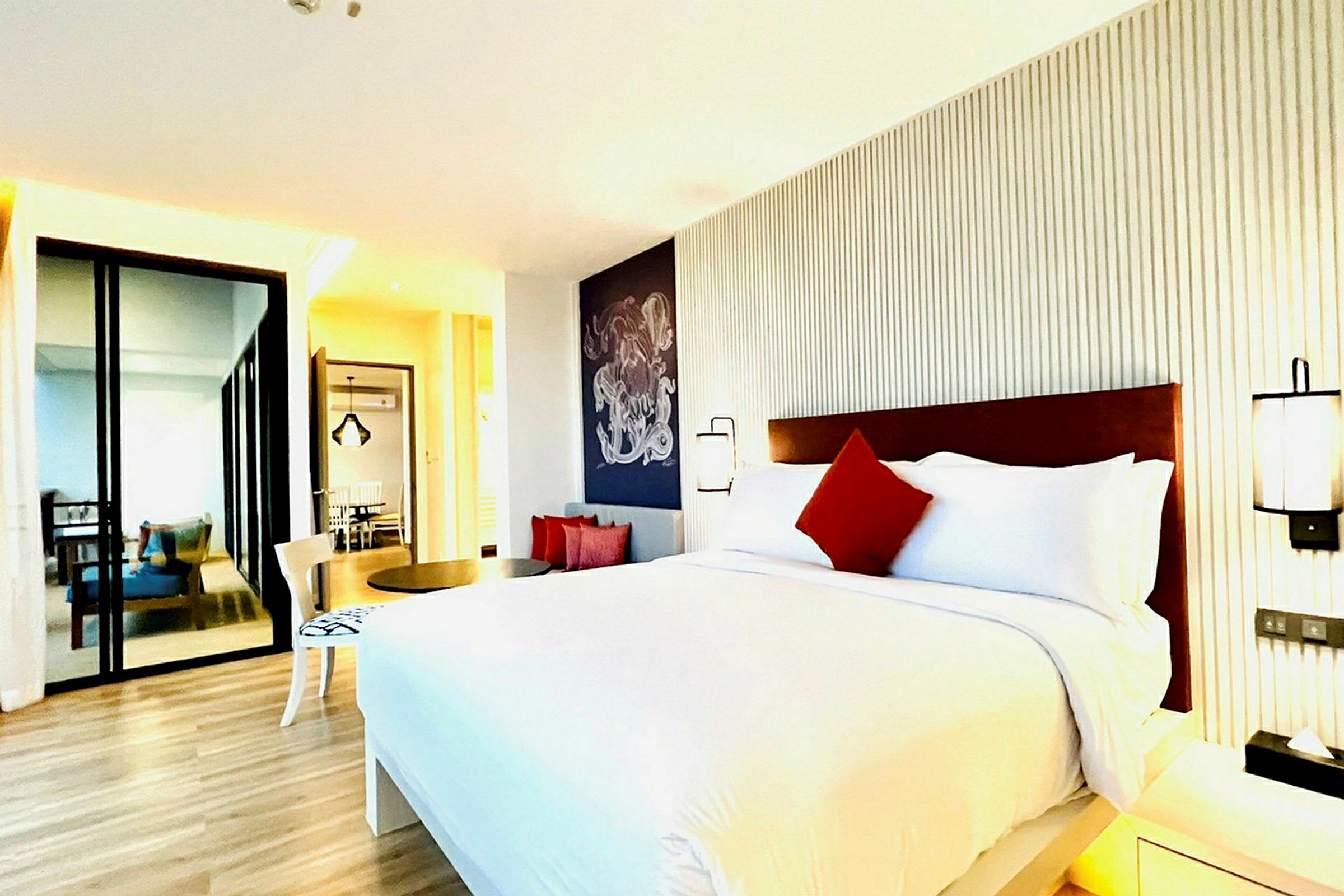 Moderni hotellihuone, jossa iso sänky, valkoiset ja punaiset tyynyt, minimalistinen sisustus ja puulattia. (Tekoälyn luoma teksti)