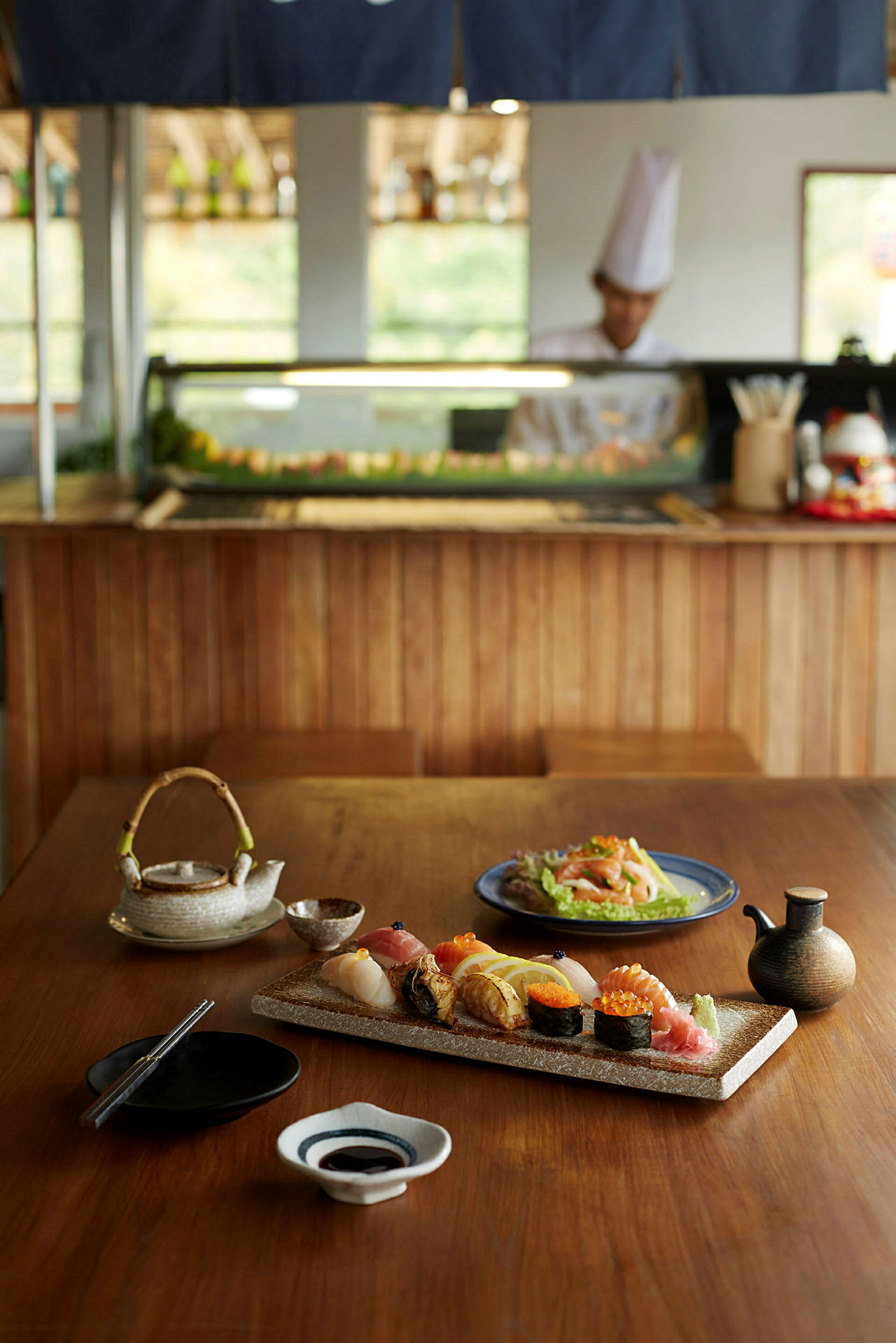 Pöydällä puualustalla sushiannos ja muita japanilaisia ruokia sekä teekannu ja lisukkeita. (Tekoälyn luoma teksti)