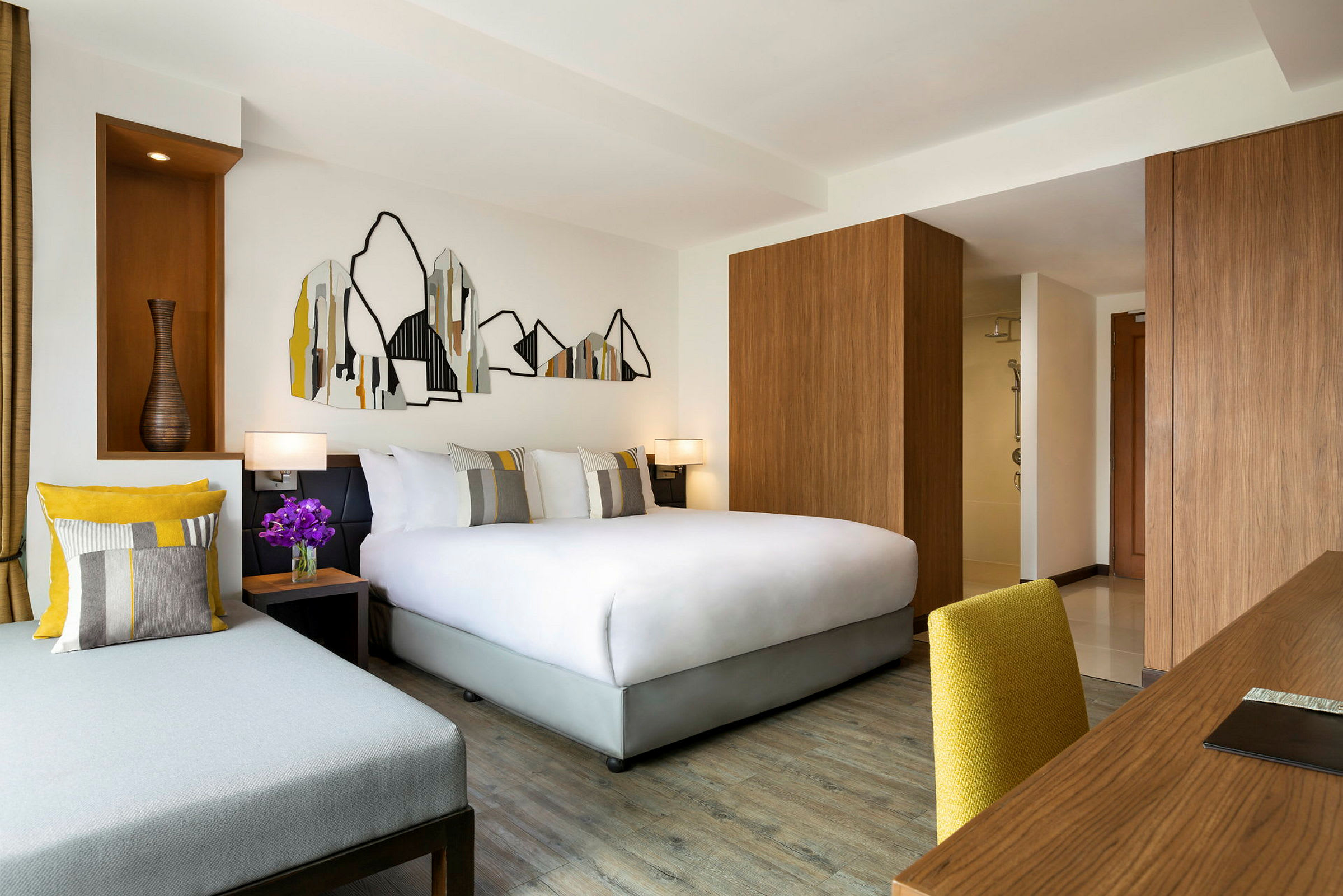Moderni hotellihuone, jossa on parivuode, sohva ja kirjoituspöytä keltaisella tuolilla. Seinällä abstrakti vuorimaisemakoriste. (Tekoälyn luoma teksti)