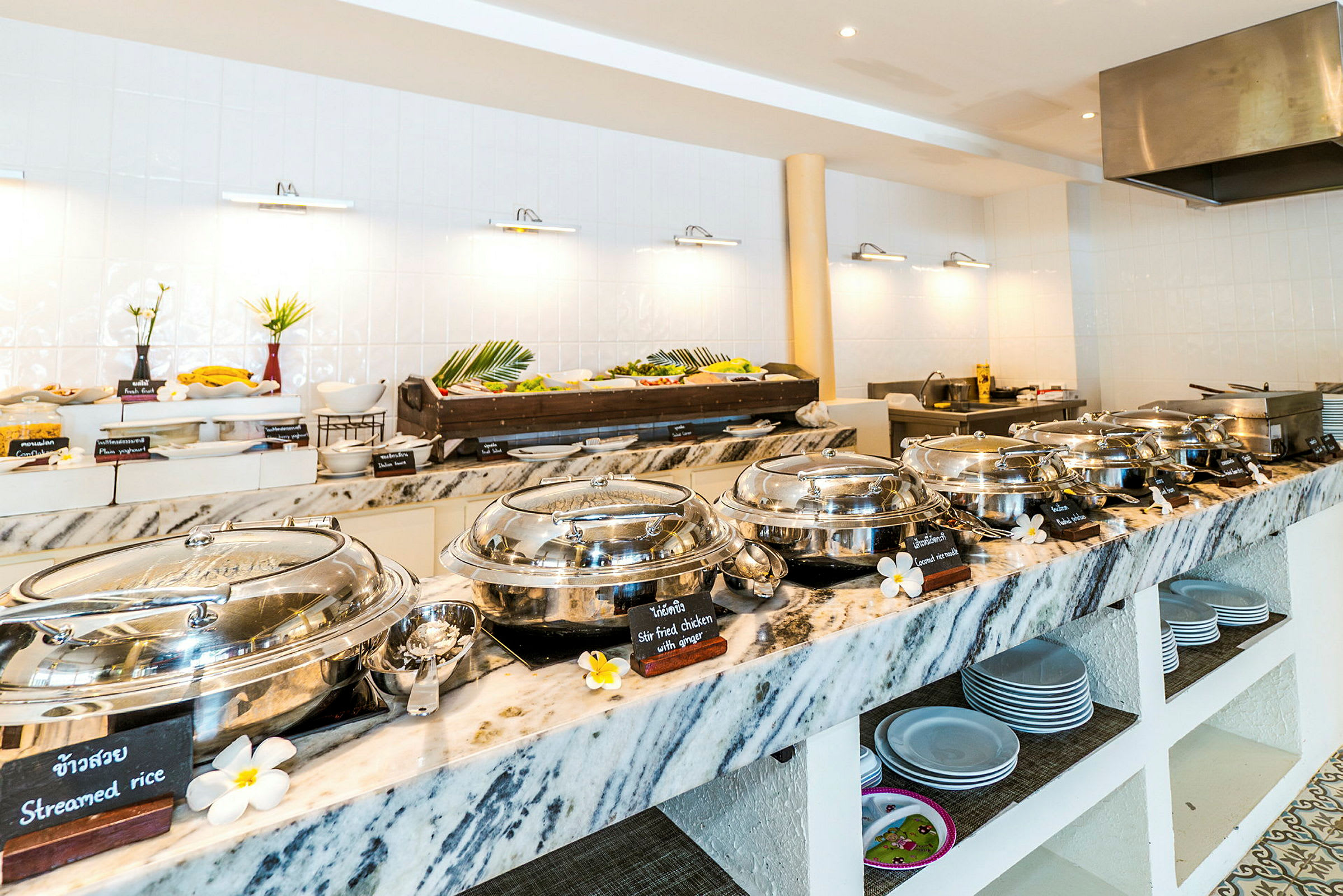 Buffet-asetelma lämpimien ruokien metalliastiat ja taustalla tarjoillut kylmät lisukkeet, ympäröitynä vaalealla sisustuksella. (Tekoälyn luoma teksti)