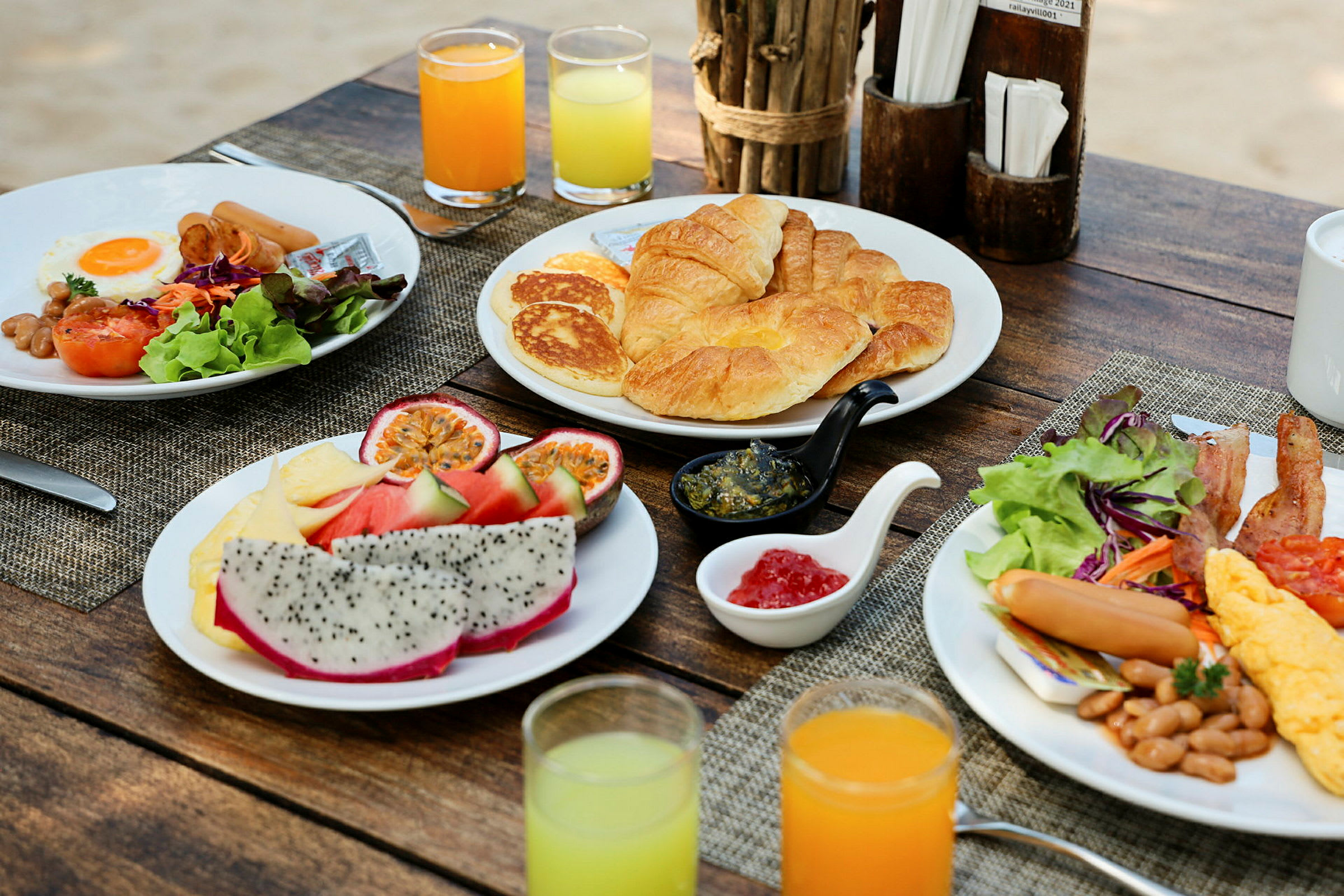 Aamiainen, jossa on erilaisia ruokia, tarjottuna rustiikkisella puupöydällä, sisältäen croissanteja, pannukakkuja, hedelmiä, munia ja salaattia, yhdessä mehulasien kanssa. (Tekoälyn luoma teksti)