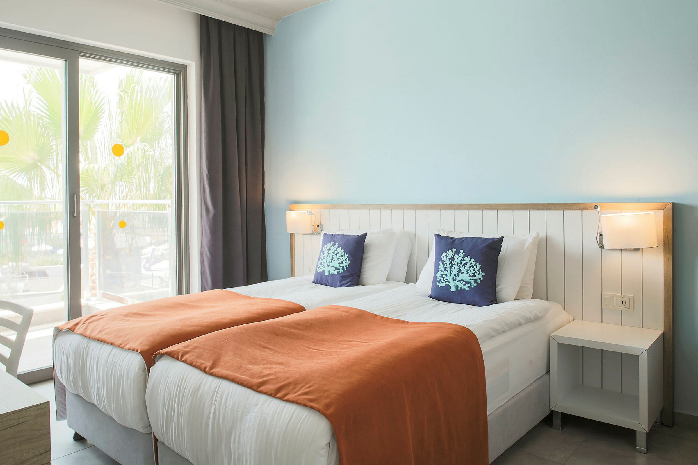 Hotellihuone, jossa on kaksi yhden hengen vuodetta, oranssit peitot ja koristeelliset tyynyt. (Tekoälyn luoma teksti)