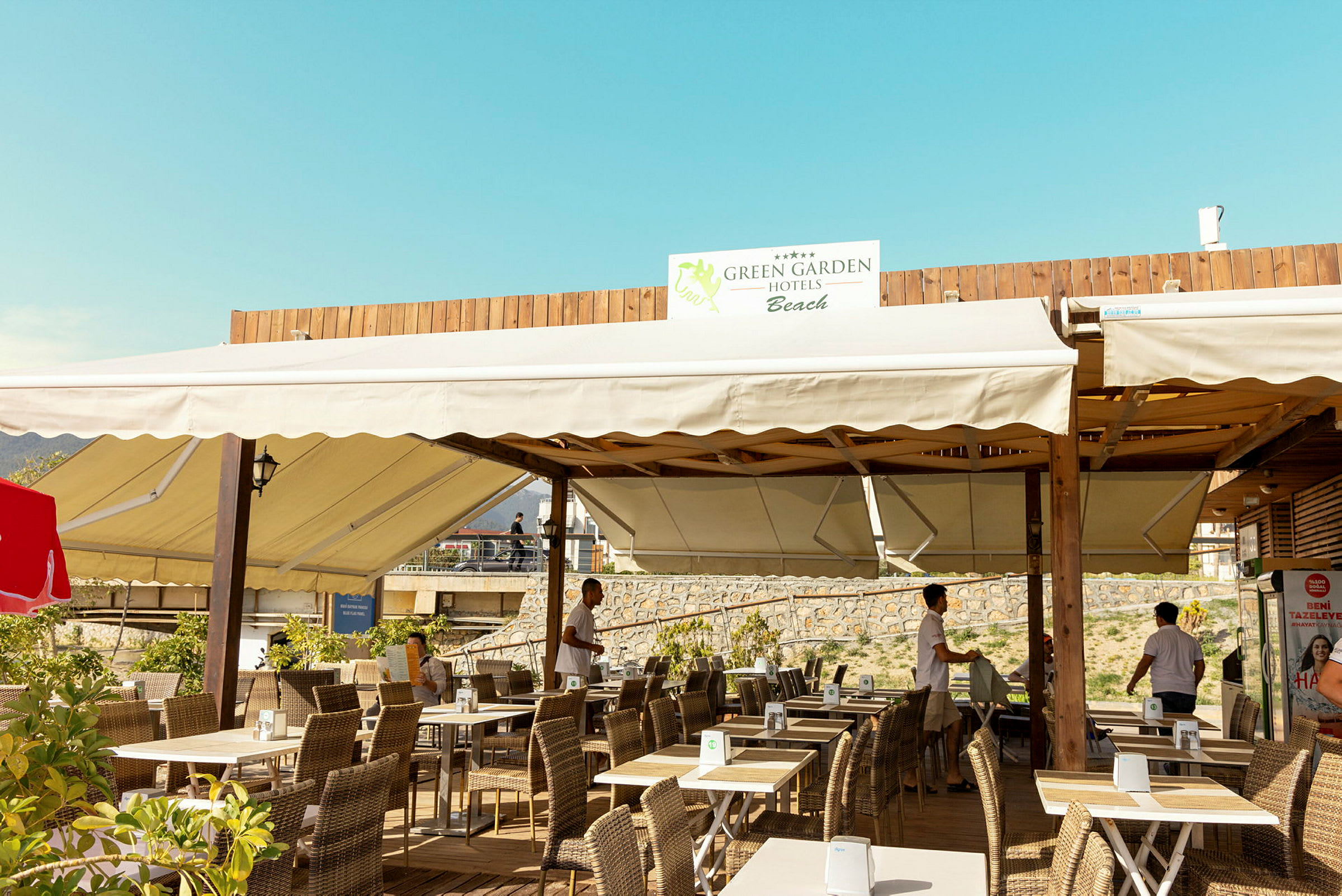 Ulkoilmaterassi, jossa on pöytiä ja tuoleja, yläpuolella oleva purjekatos ja kyltti, jossa lukee 'Green Garden Hotels Beach'. (Tekoälyn luoma teksti)