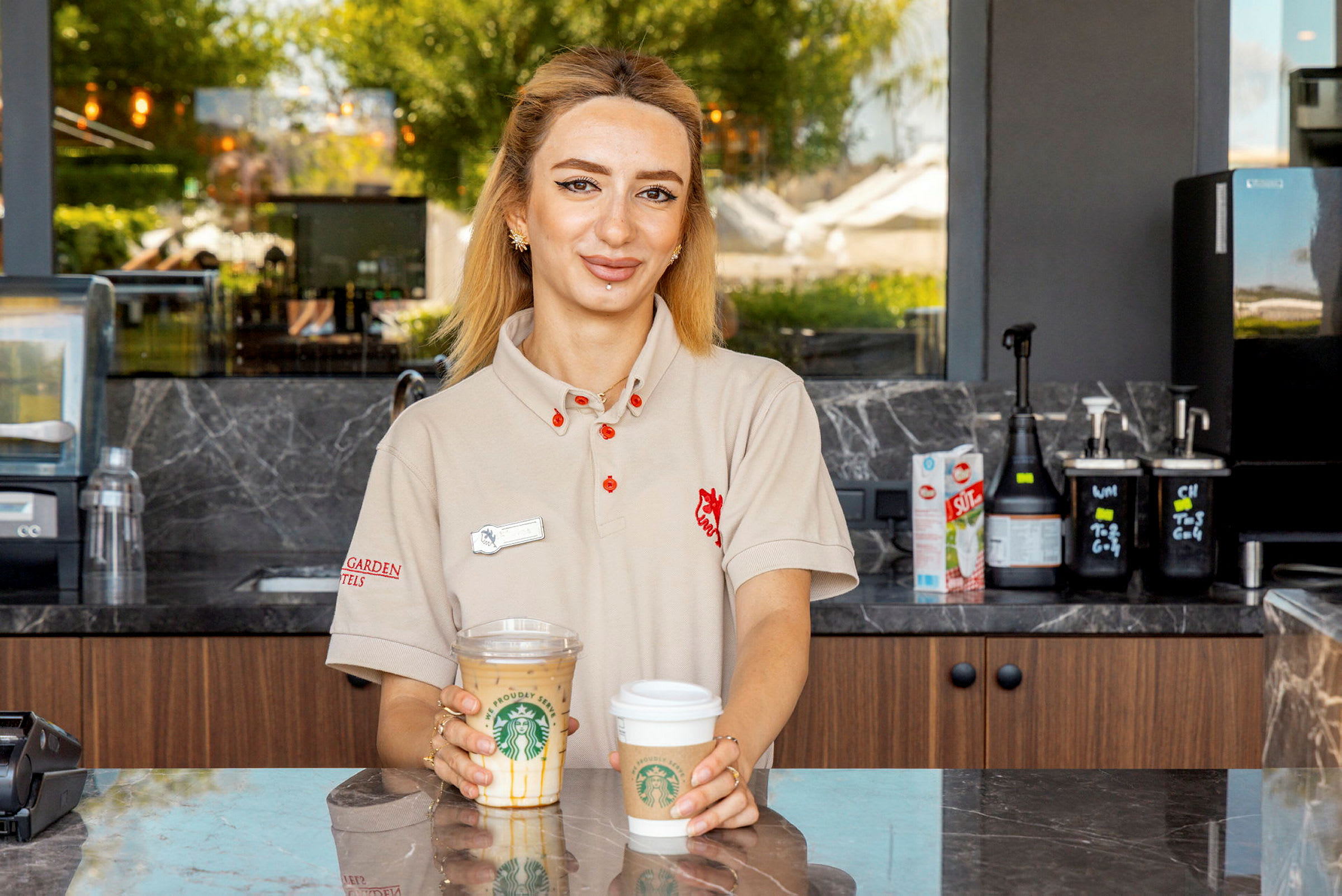 Nainen seisoo tiskin takana ja pitää kahta Starbucks-kahvikuppia käsissään. (Tekoälyn luoma teksti)
