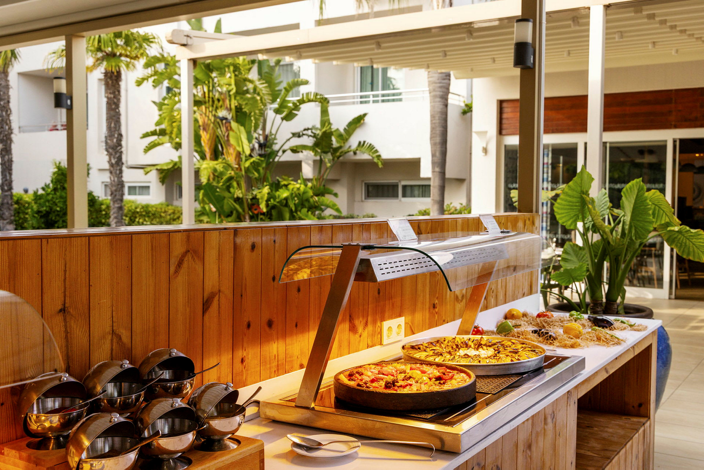 Buffet, jossa ruokaa, mukaan lukien kaksi pizzaa, jota ympäröivät vihreät kasvit ja ulkoilmaympäristö. (Tekoälyn luoma teksti)