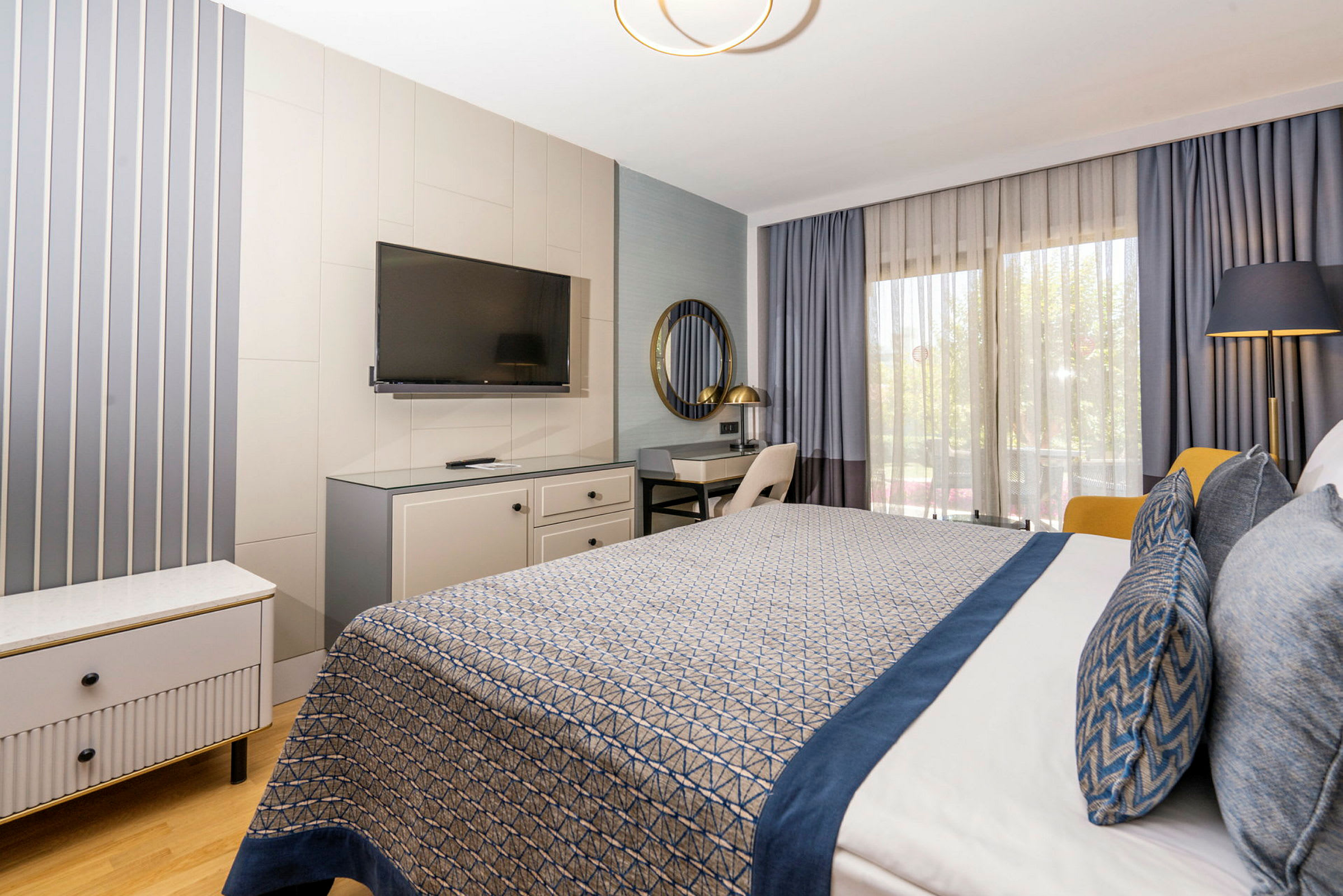 Hotellihuone, jossa moderni sisustus, mukaan lukien sinisillä yksityiskohdilla varustettu sänky, työpöytä, televisio ja suuret ikkunat verhoilla. (Tekoälyn luoma teksti)