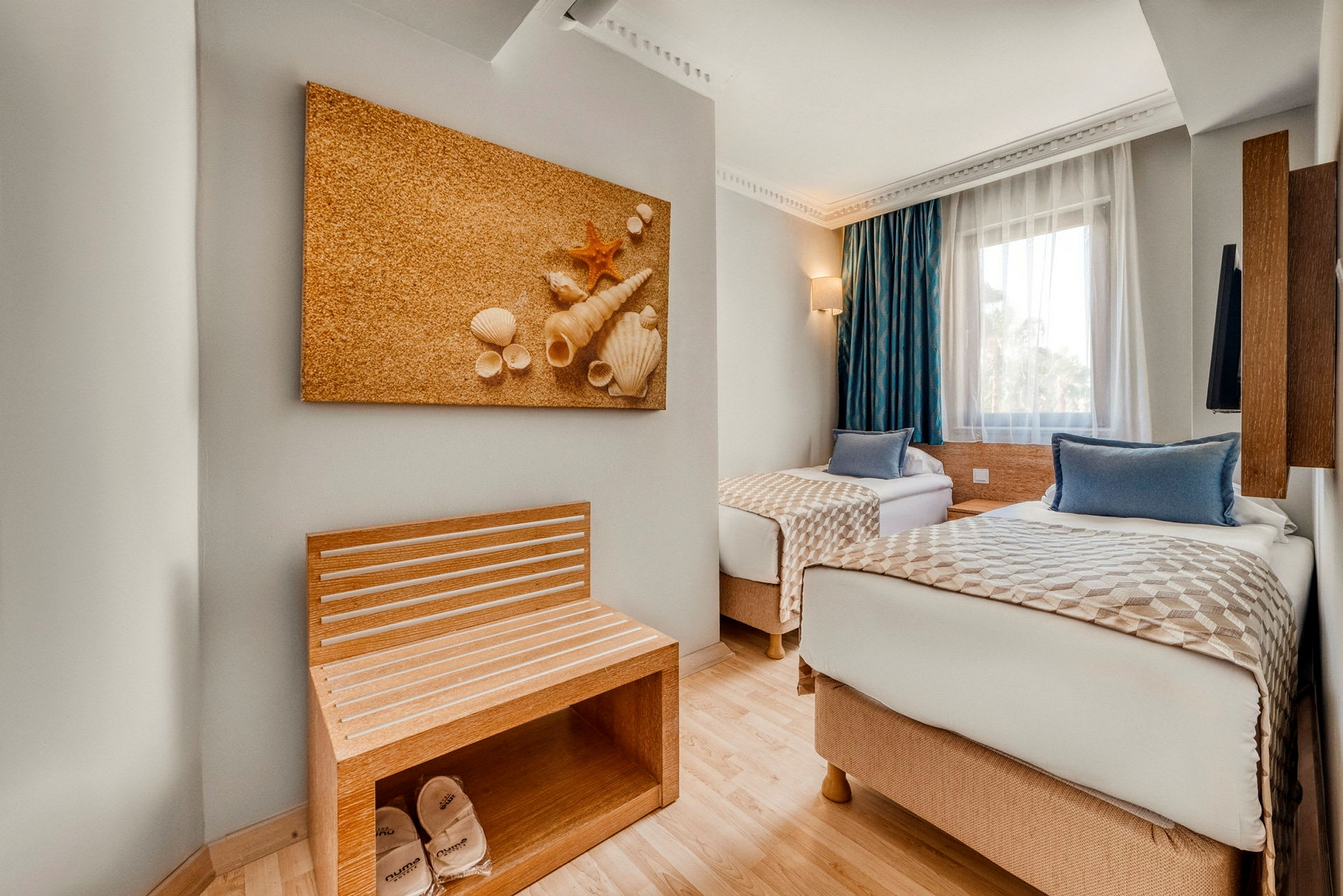 Kaksi yhden hengen vuodetta hotellihuoneessa, jossa on simpukkakoriste seinällä ja penkki, jossa on tohveleita. Vieressä on ikkuna sinisillä verhoilla. (Tekoälyn luoma teksti)
