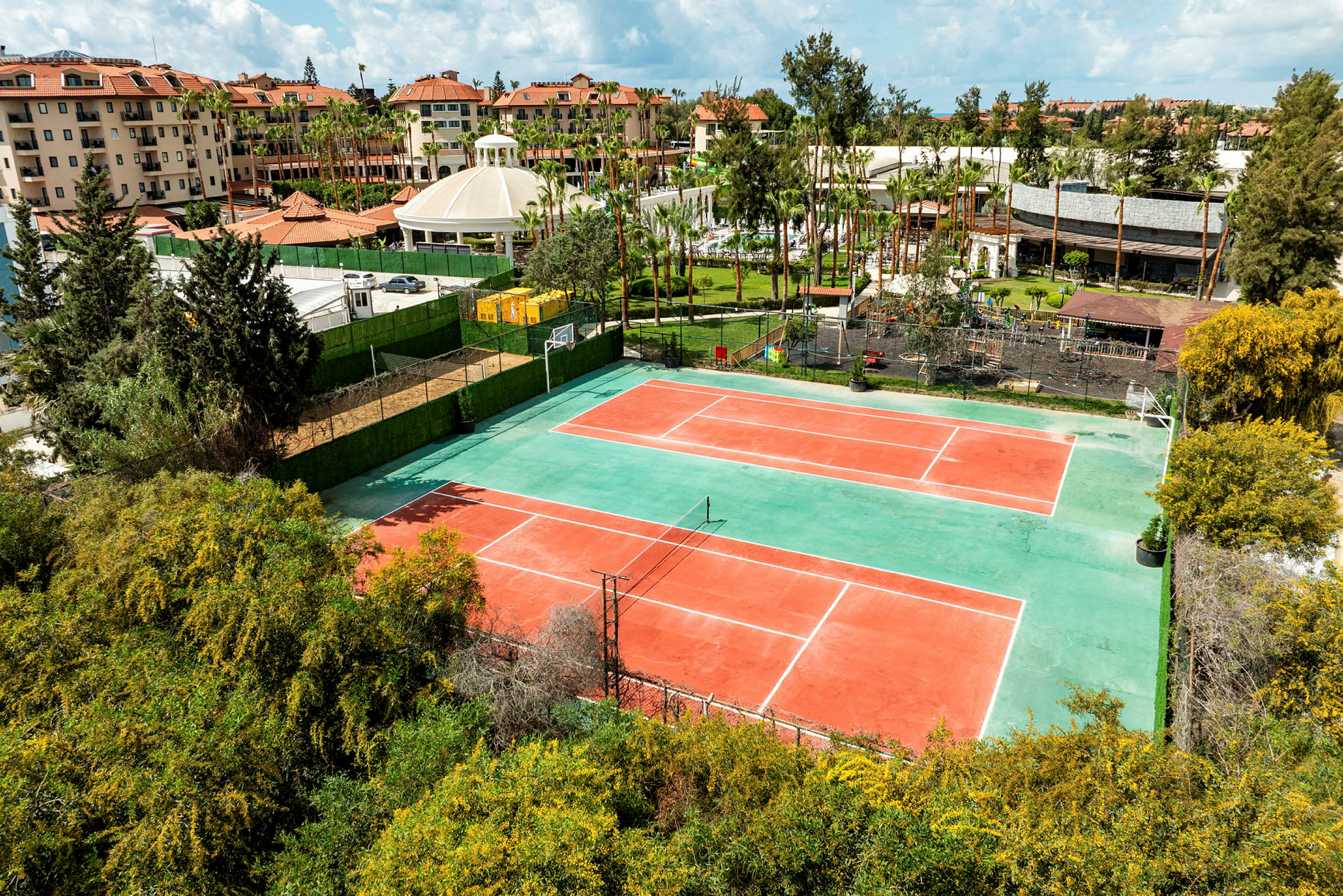 Kuva kahdesta tenniskentästä, joita ympäröivät viheralueet ja taustalla näkyvät rakennukset. (Tekoälyn luoma teksti)