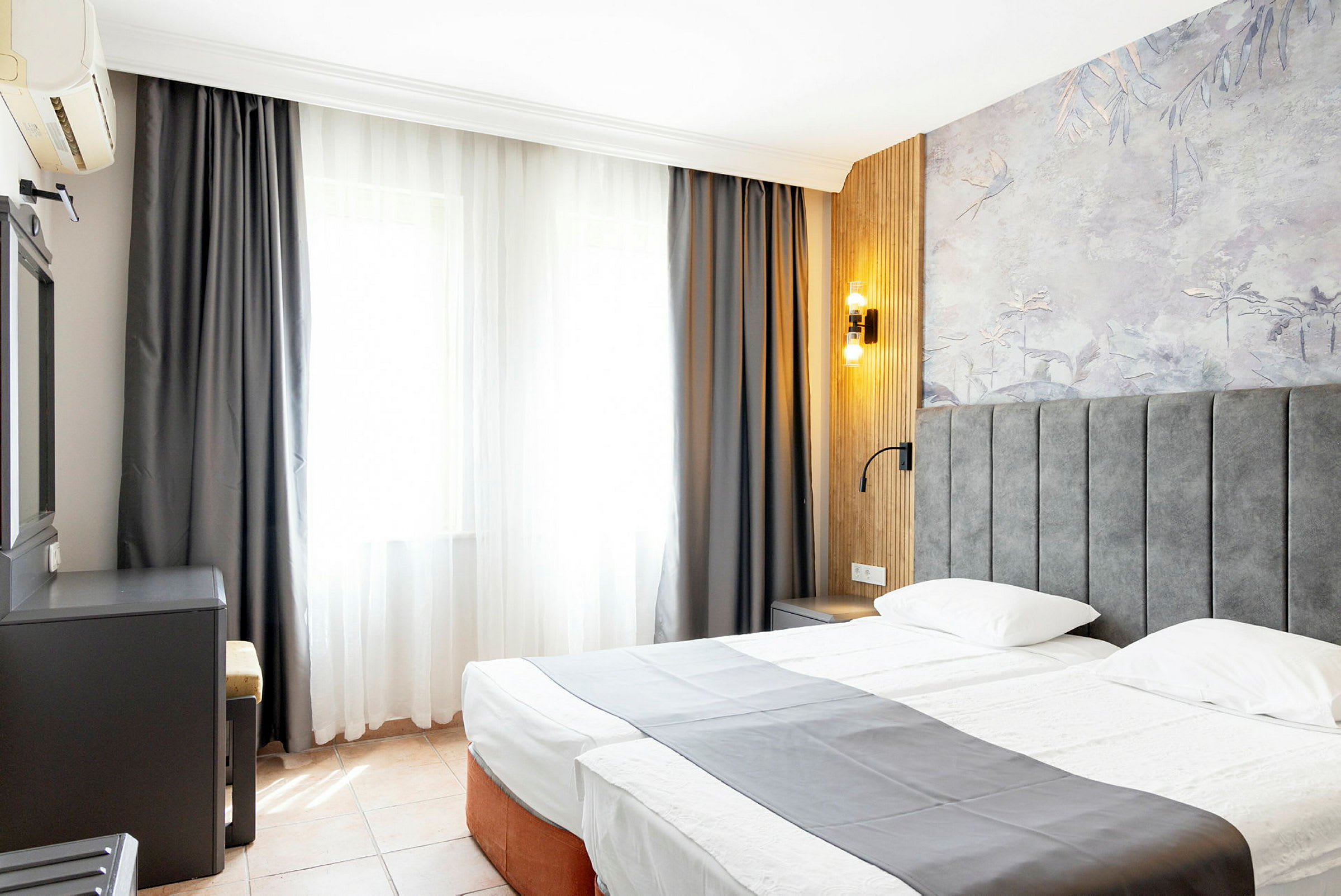Hotellihuone, jossa on parivuode, harmaa päiväpeitto, ikkuna vaaleilla verhoilla ja seinäpaneli koristeellisin kuvioin. (Tekoälyn luoma teksti)