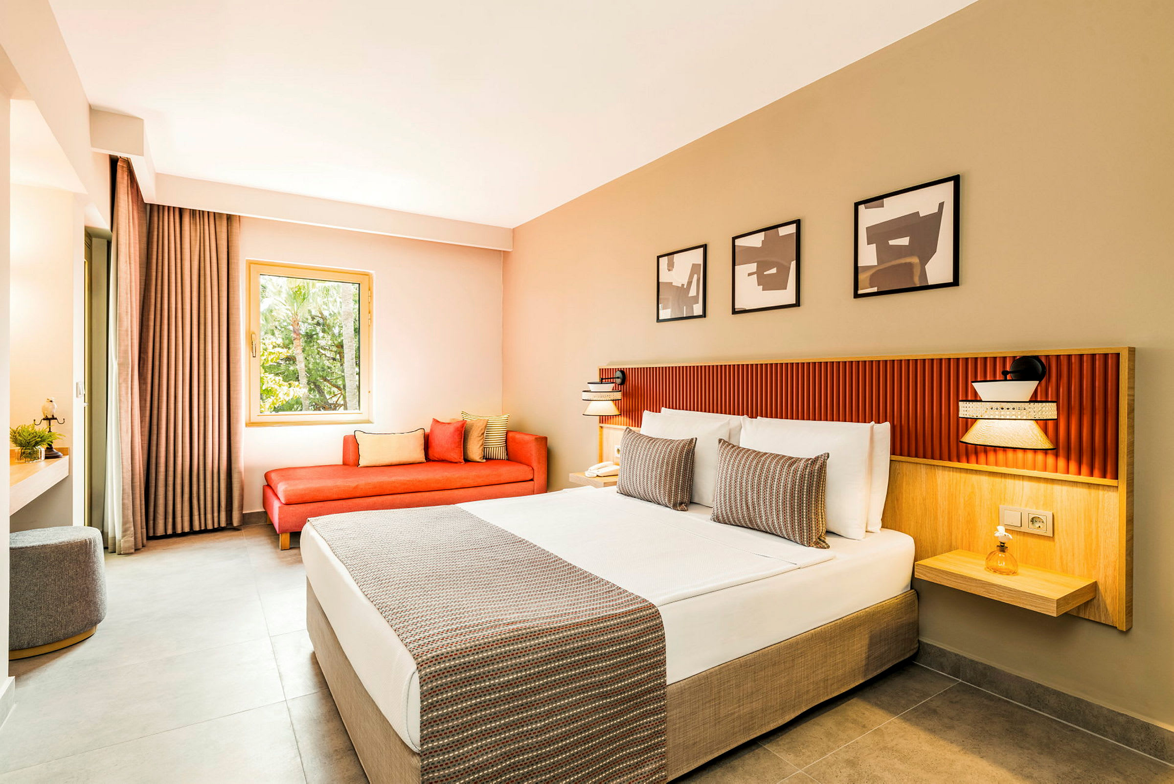 Moderni hotellihuone, jossa leveä sänky valkoisin lakanoilla ja koristeellisilla tyynyillä. Ikkunan vieressä on punainen sohva ja kolme taideteosta sängyn yllä seinällä. (Tekoälyn luoma teksti)