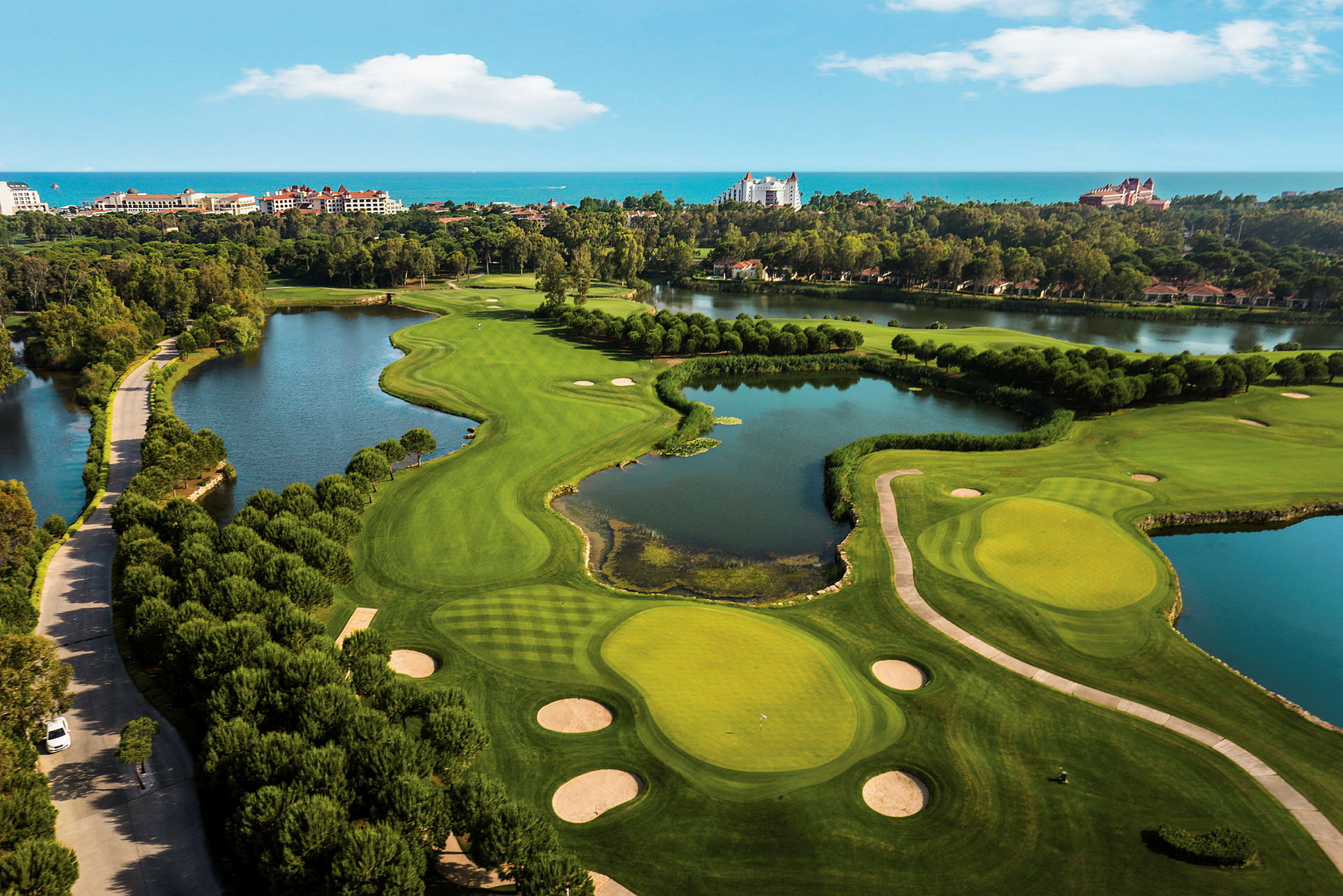 Ilmakuva golfkentästä, jossa vihreät väylyt, bunkkerit ja vesiesiintymät, ympäröitynä hotelleilla ja puilla. (Tekoälyn luoma teksti)