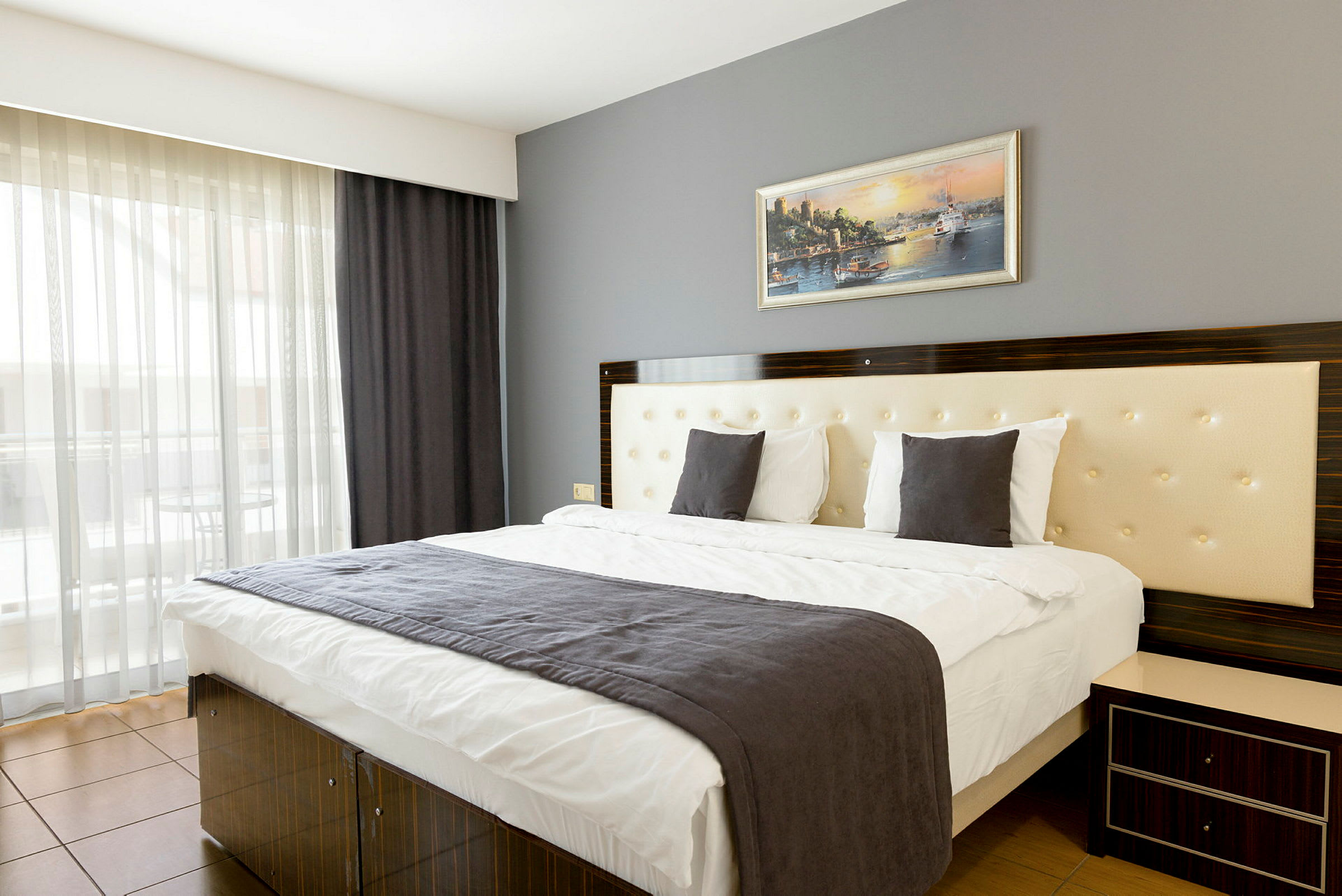Moderni hotellihuone, jossa kaksoisvuode, harmaa päiväpeite ja seinällä oleva taulu. (Tekoälyn luoma teksti)