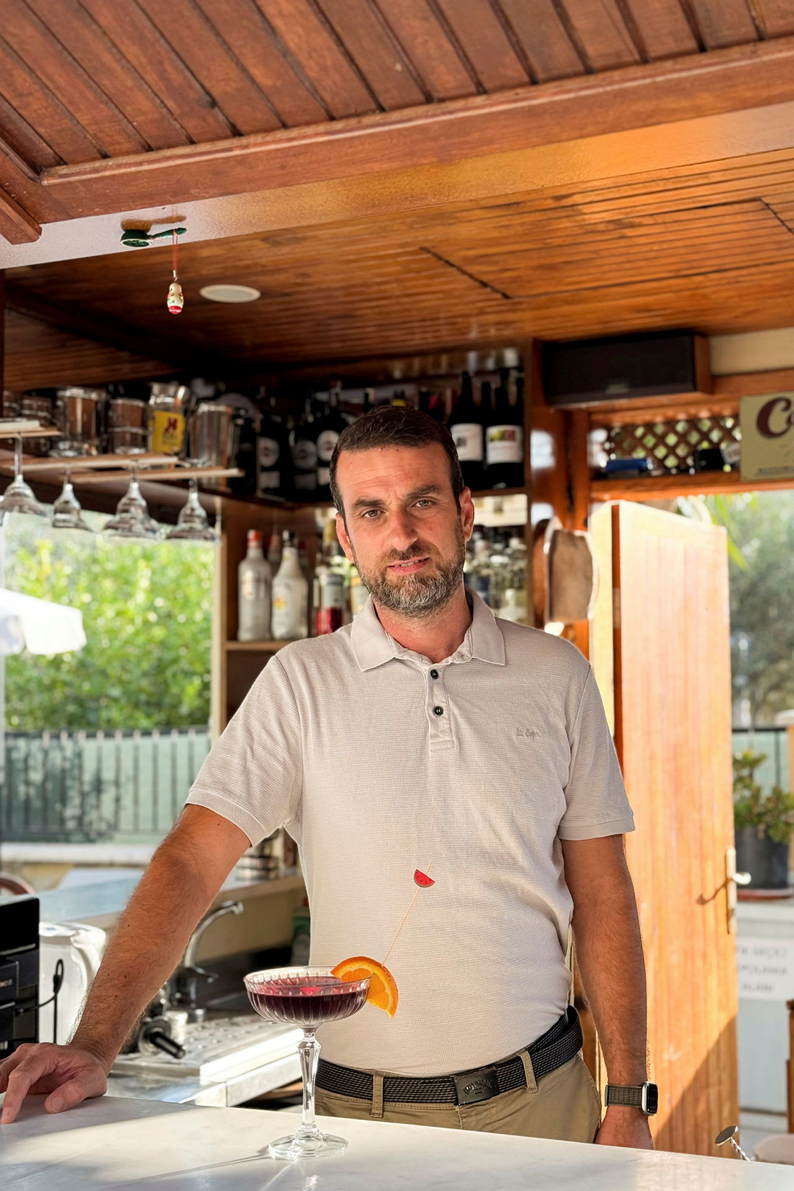 Valkoinen paita baarissa, taustalla hyllyjä täynnä pulloja, edessä cocktail-lasi värillisellä juomalla. (Tekoälyn luoma teksti)