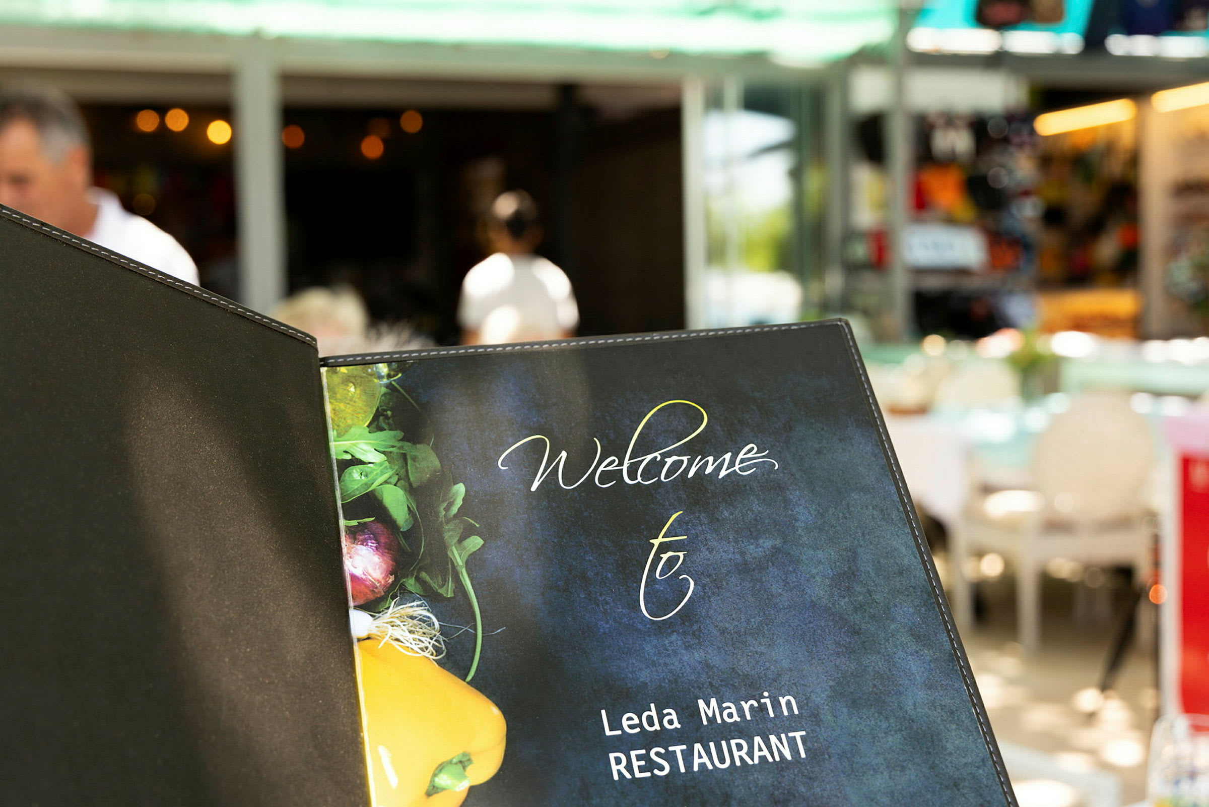 Ravintolamenun kansi, jossa lukee 'Welcome to Leda Marin RESTAURANT' ja kasvisten kuva etuosassa. (Tekoälyn luoma teksti)