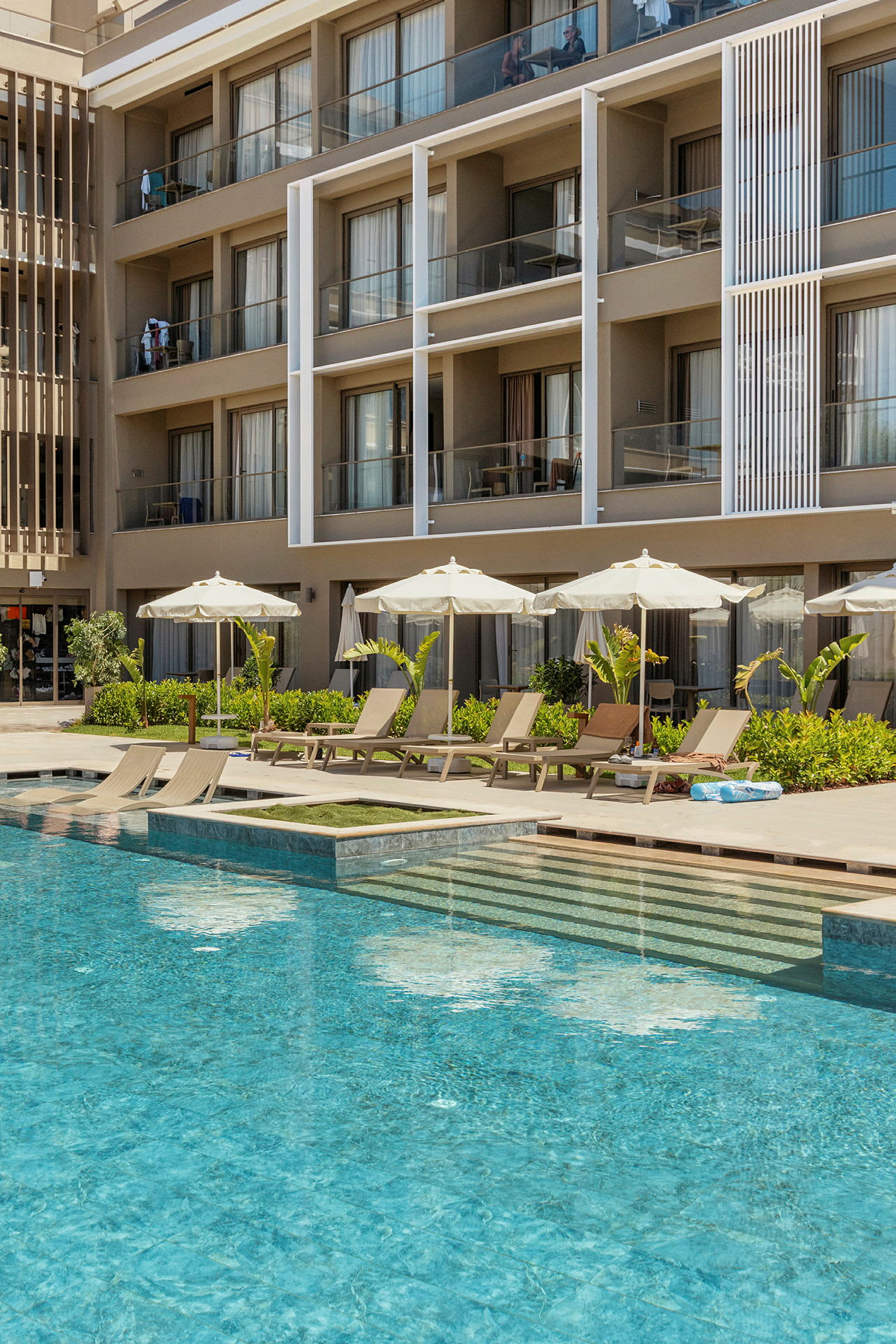 Hotellin julkisivu parvekkeineen ja uima-allas, jossa on aurinkotuoleja ja varjoja. (Tekoälyn luoma teksti)