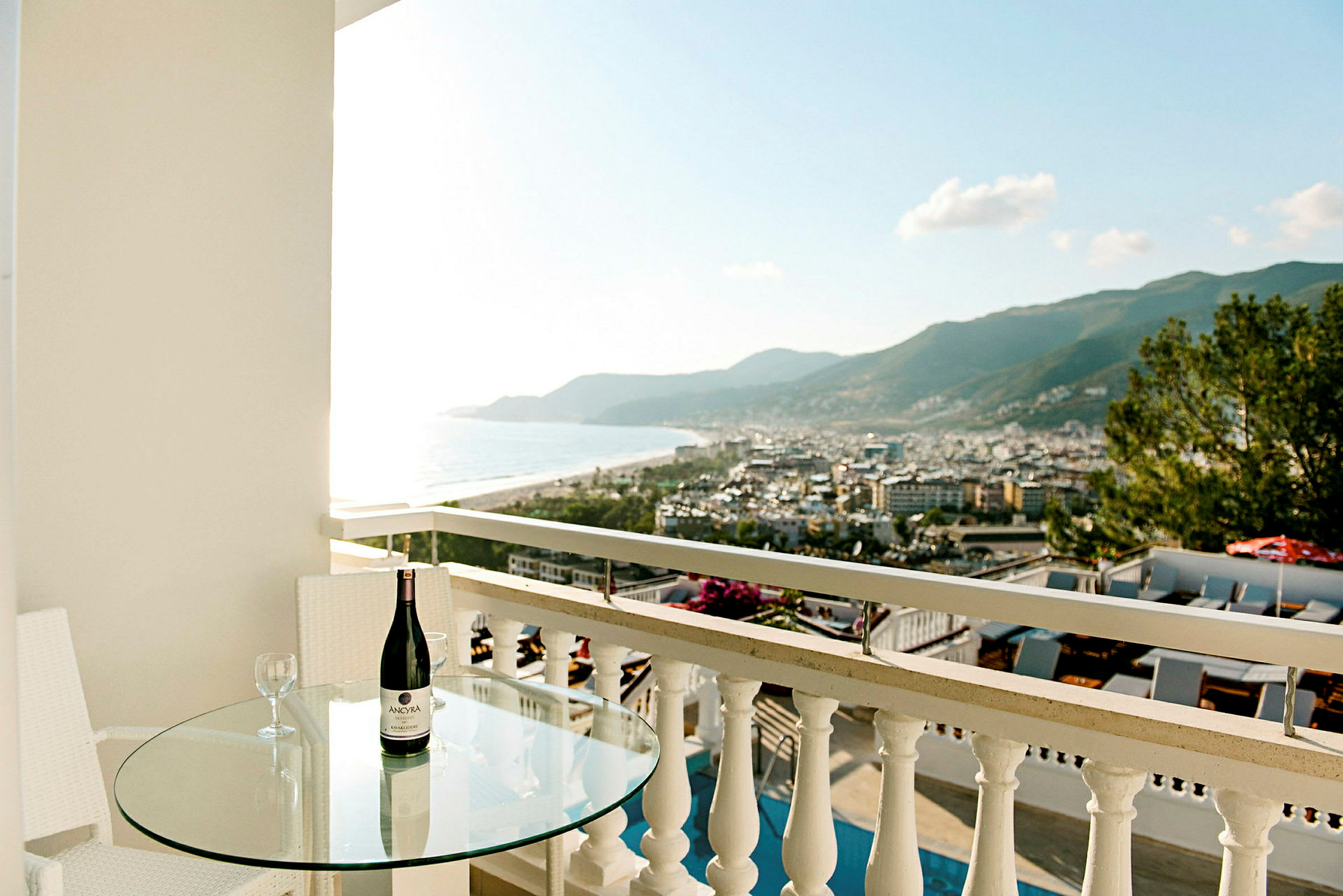 Parveke, jossa lasipöytä, viinipullo ja näkymä kaupungin ja rannikolle. (Tekoälyn luoma teksti)