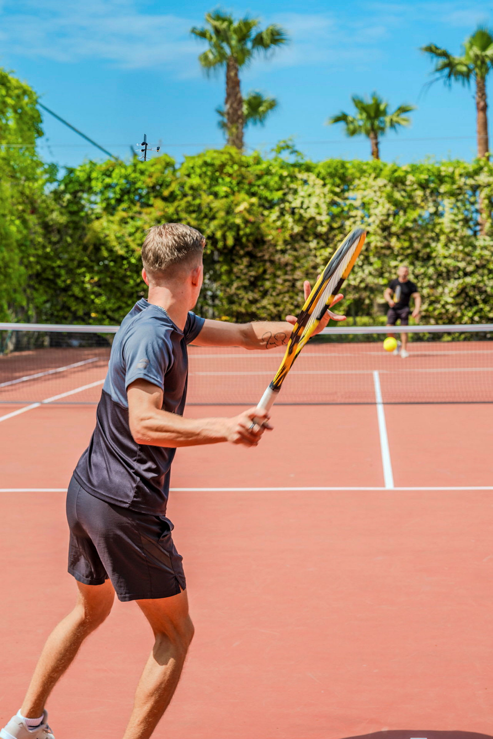 Kaksi henkilöä pelaa tennistä ulkona tenniskentällä, jota ympäröi vehreys ja palmut. (Tekoälyn luoma teksti)