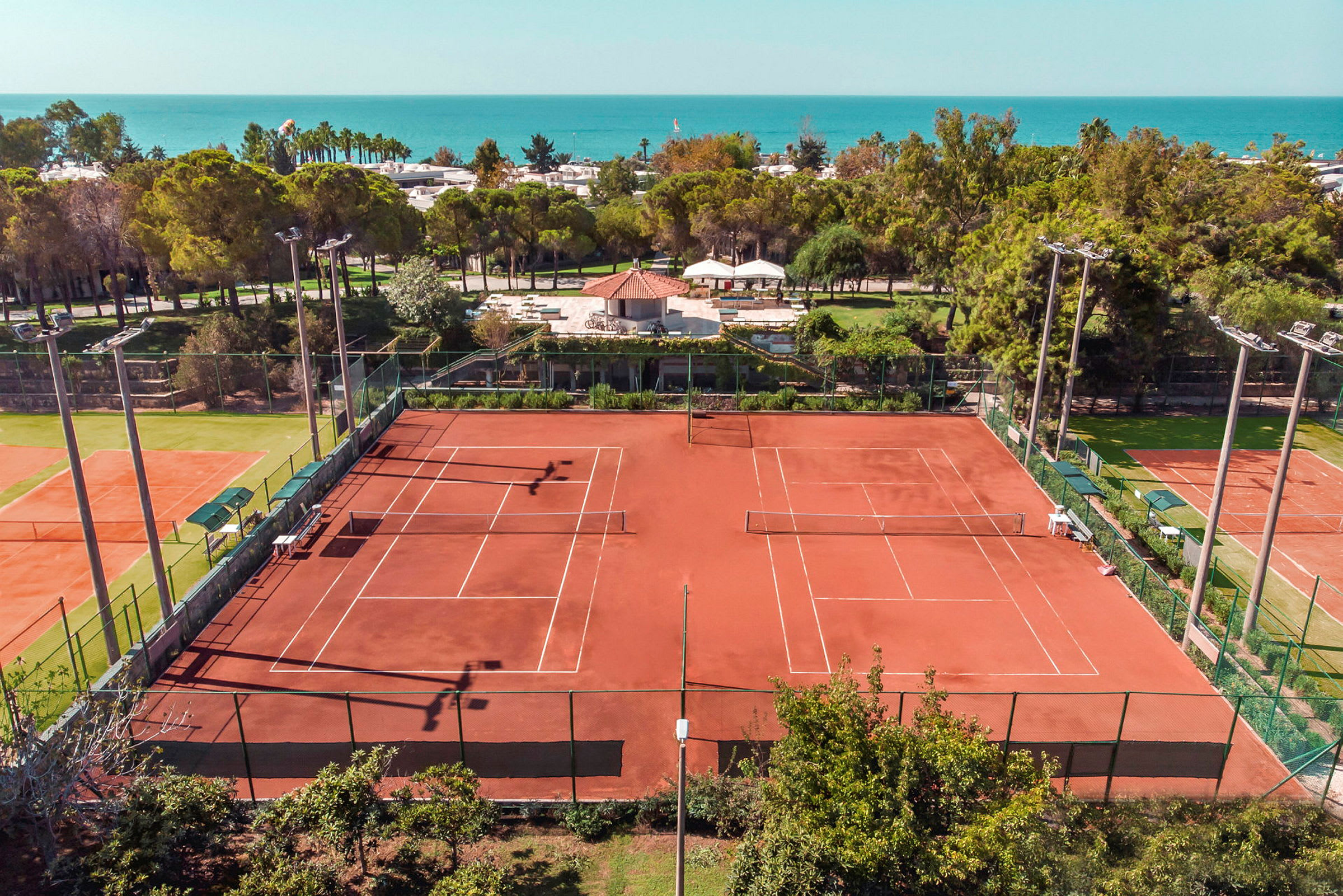 Kuva kahdesta punaisen hiekan tenniskentästä vehreän kasvillisuuden keskellä ja merinäköalalla. (Tekoälyn luoma teksti)