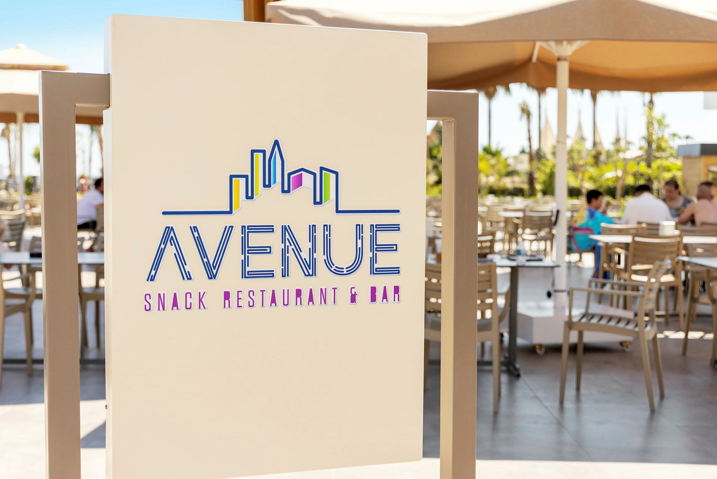Kyltti, jossa lukee "Avenue Snack Restaurant & Bar" ja tyylitelty siluettikuva kaupunkimaisemasta. Taustalla näkyy terassilla olevia pöytiä ja tuoleja varjojen alla. (Tekoälyn luoma teksti)