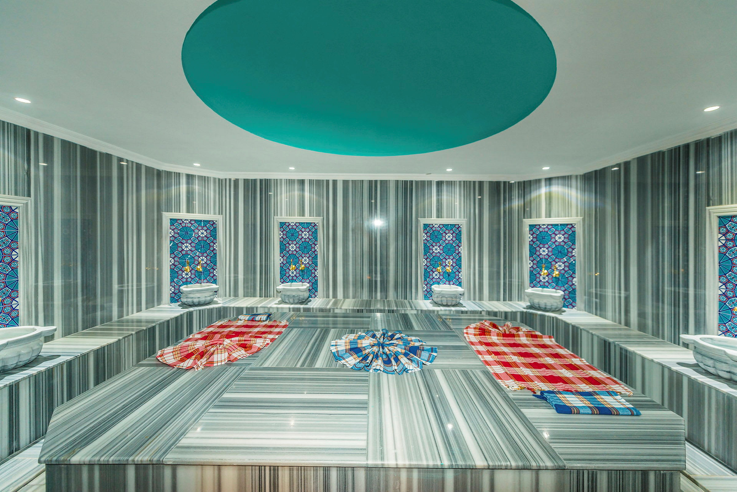 Hamam-huone, jossa on marmoripenkkejä, värikkäitä pyyhkeitä ja koristeellisia mosaiikki-ikkunoita. (Tekoälyn luoma teksti)