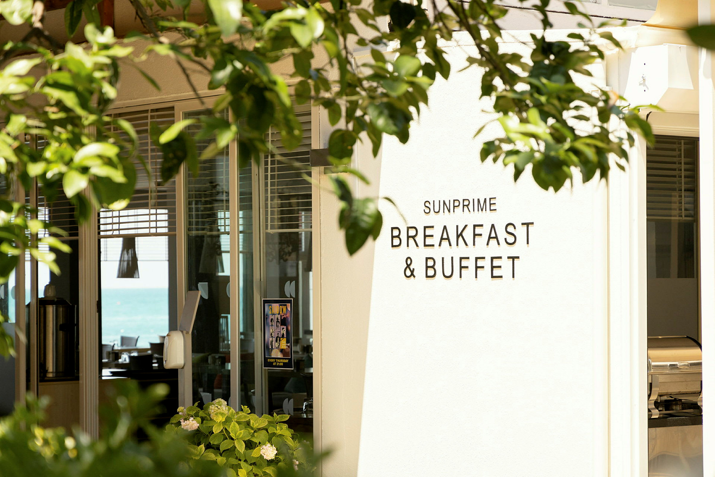 Kilpi, jossa lukee 'Sunprime Breakfast & Buffet', ravintolan sisäänkäynnillä, vehreyden ympäröimänä. (Tekoälyn luoma teksti)