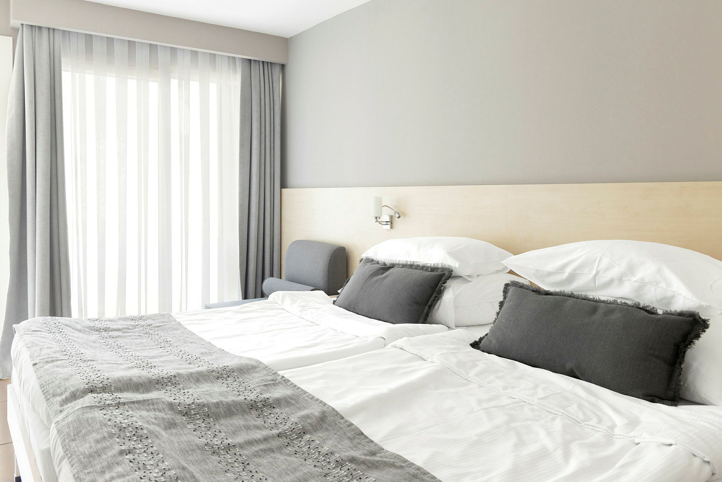 Hotellihuone, jossa on parivuode, vaaleat lakanat ja harmaat koristeelliset tyynyt. (Tekoälyn luoma teksti)
