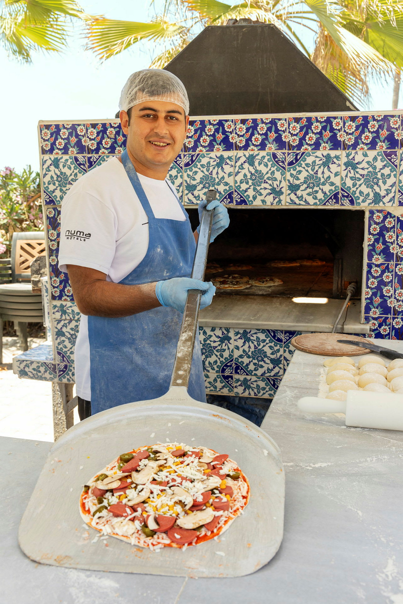 Pizzakokki pitelee lapioita, jossa on valmiiksi valmistettu pizza, sinivalkoisilla kaakeleilla koristellun uunin edessä. (Tekoälyn luoma teksti)