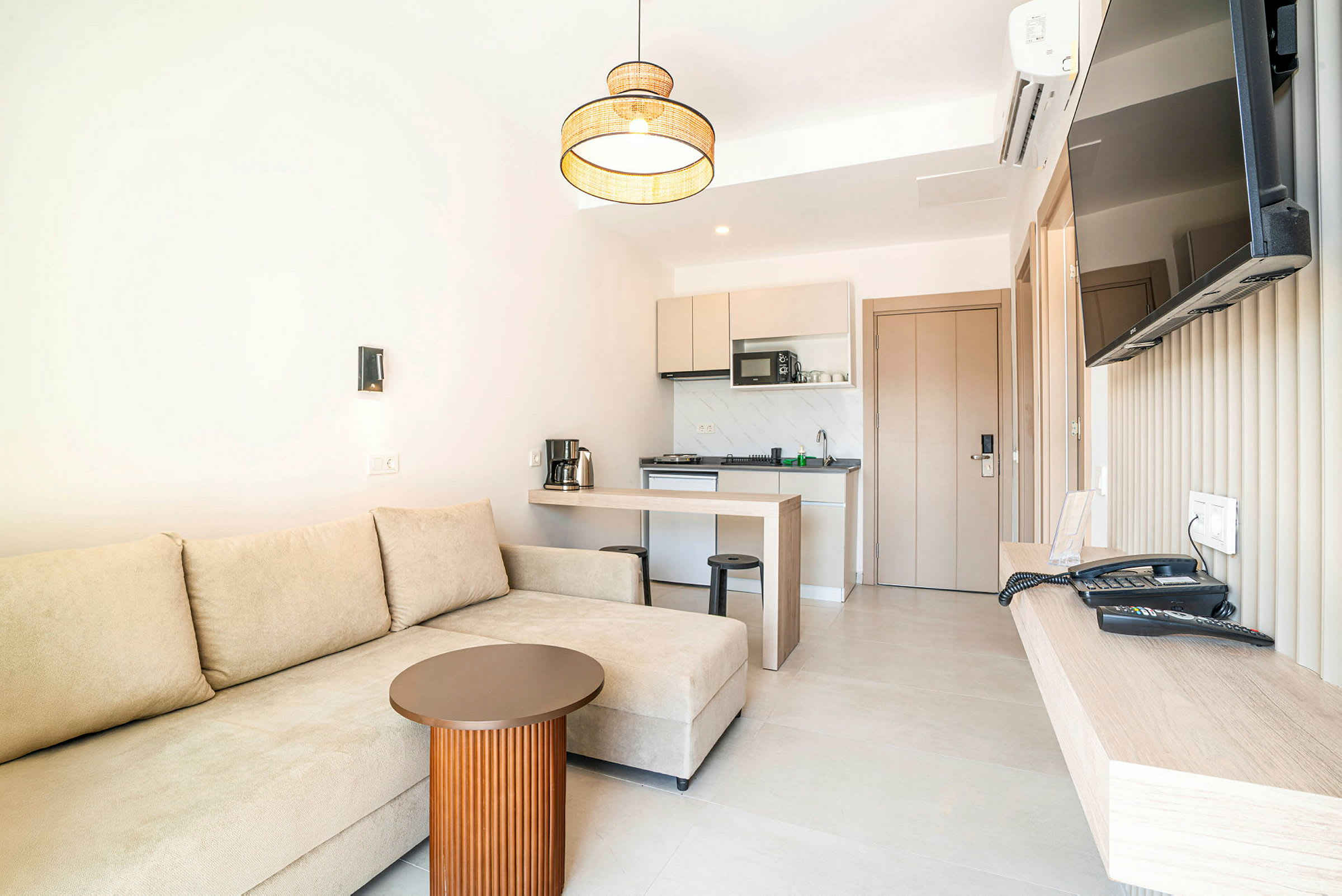 Hotellihuone, jossa on beige sohva, pieni pöytä, keittiö ja seinälle asennettu televisio. (Tekoälyn luoma teksti)