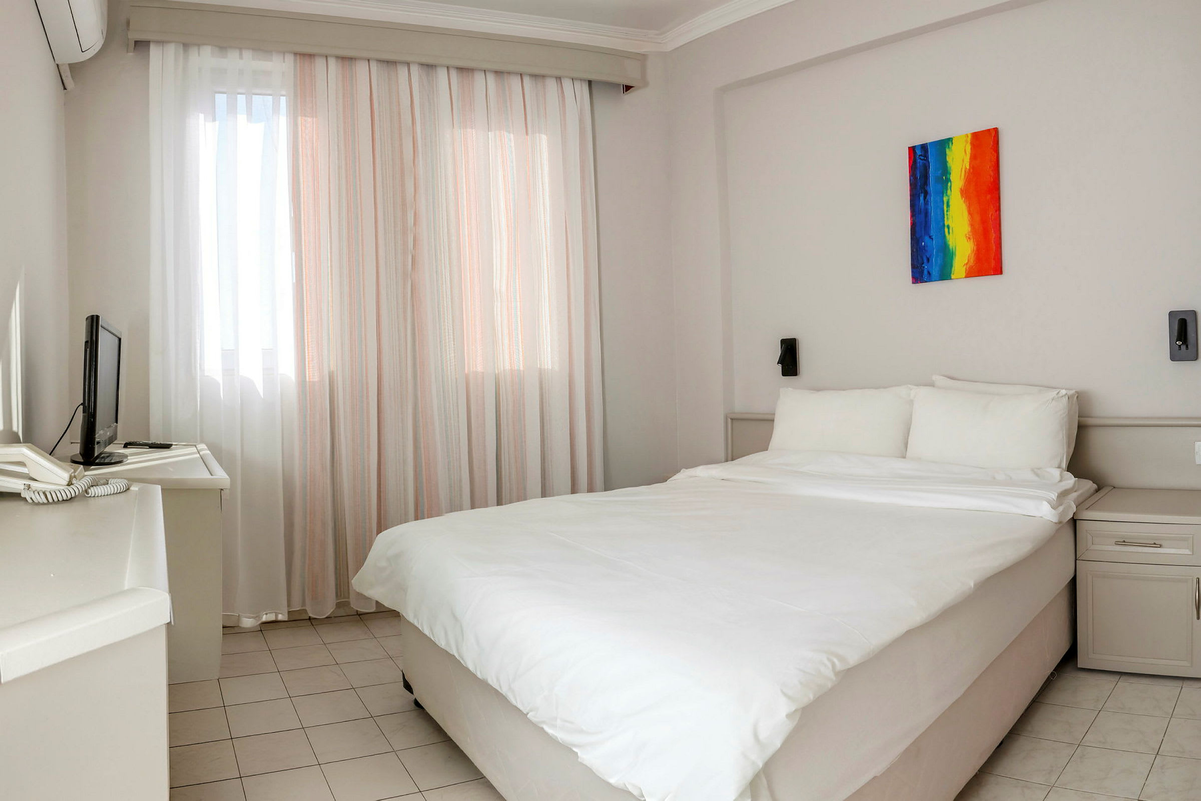 Hotellihuone, jossa on parivuode. Seinät ovat vaaleat, ja huoneessa on värikäs taideteos sängyn päädyn yläpuolella. Lähistöllä on myös pieni televisio ja kirjoituspöytä. (Tekoälyn luoma teksti)