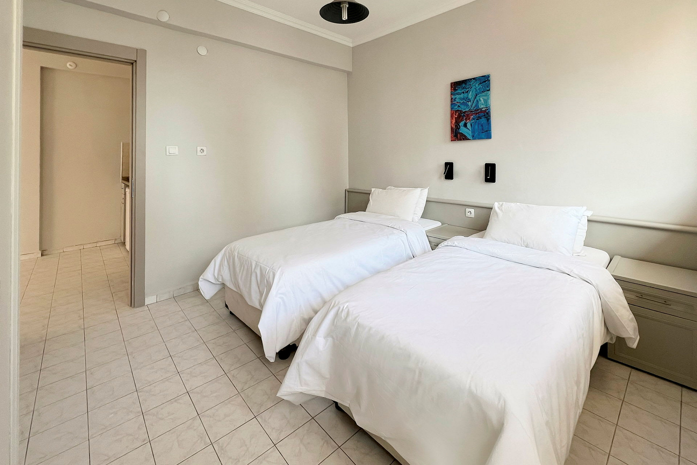 Hotellihuone, jossa on kaksi yhden hengen sänkyä, valkoiset vuodevaatteet ja vaalealaattainen lattia. Seinällä sänkyjen yläpuolella on valaisimia ja maalaus. (Tekoälyn luoma teksti)