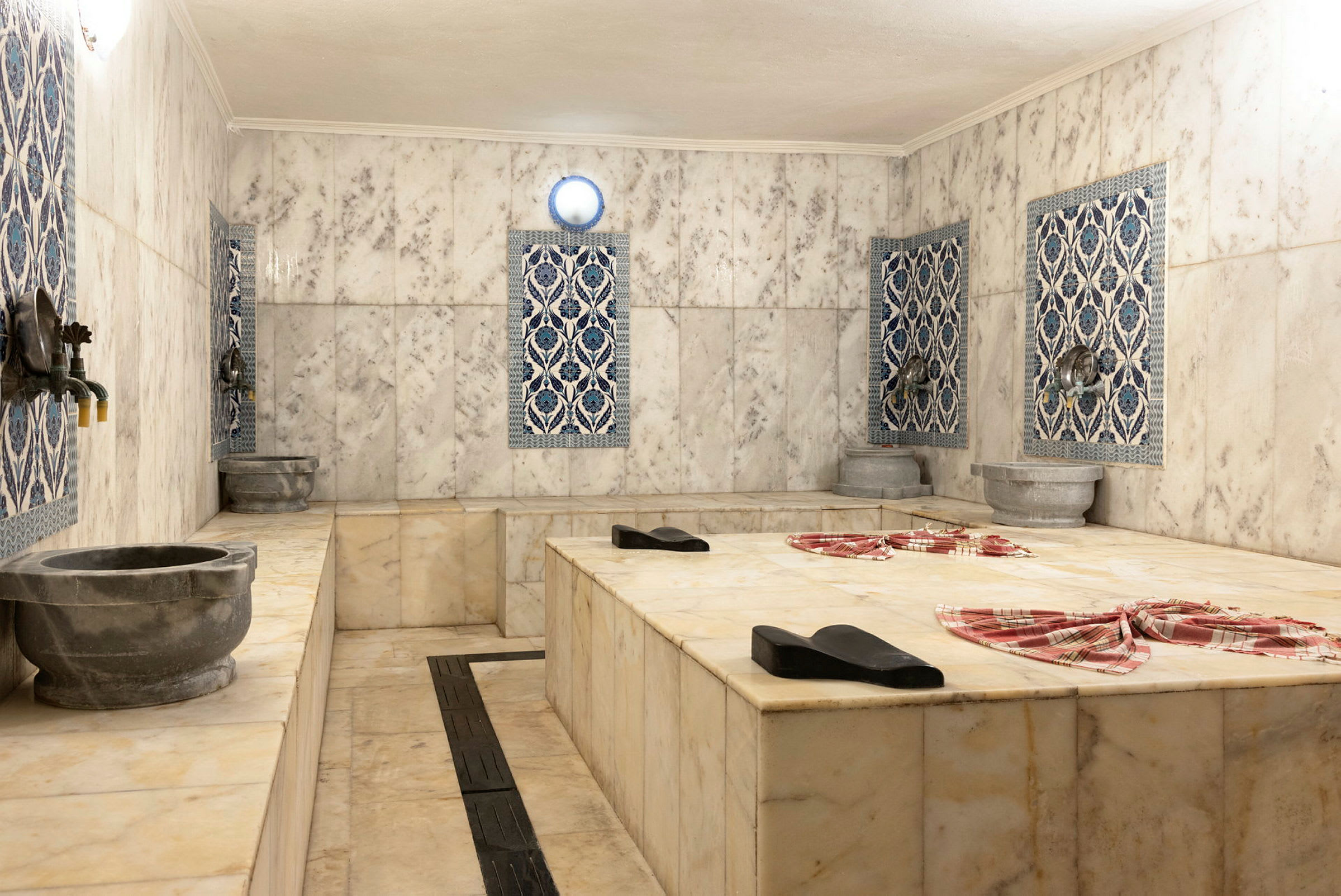 Perinteinen hamam, jossa on marmorialustoja ja koristeellisia kaakeleita seinillä. Kivisiä altaita ja hanoja näkyy ympärillä. (Tekoälyn luoma teksti)