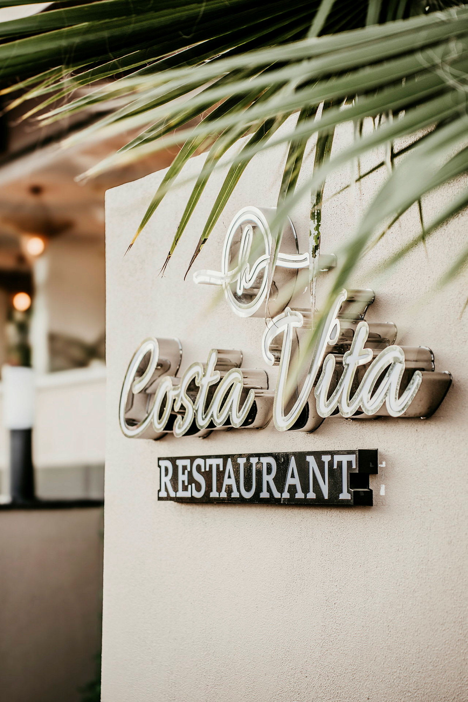 Seinässä oleva kyltti, jossa lukee 'Costa Vita Restaurant' ja jonka ympärillä on palmulehti koristeet. (Tekoälyn luoma teksti)