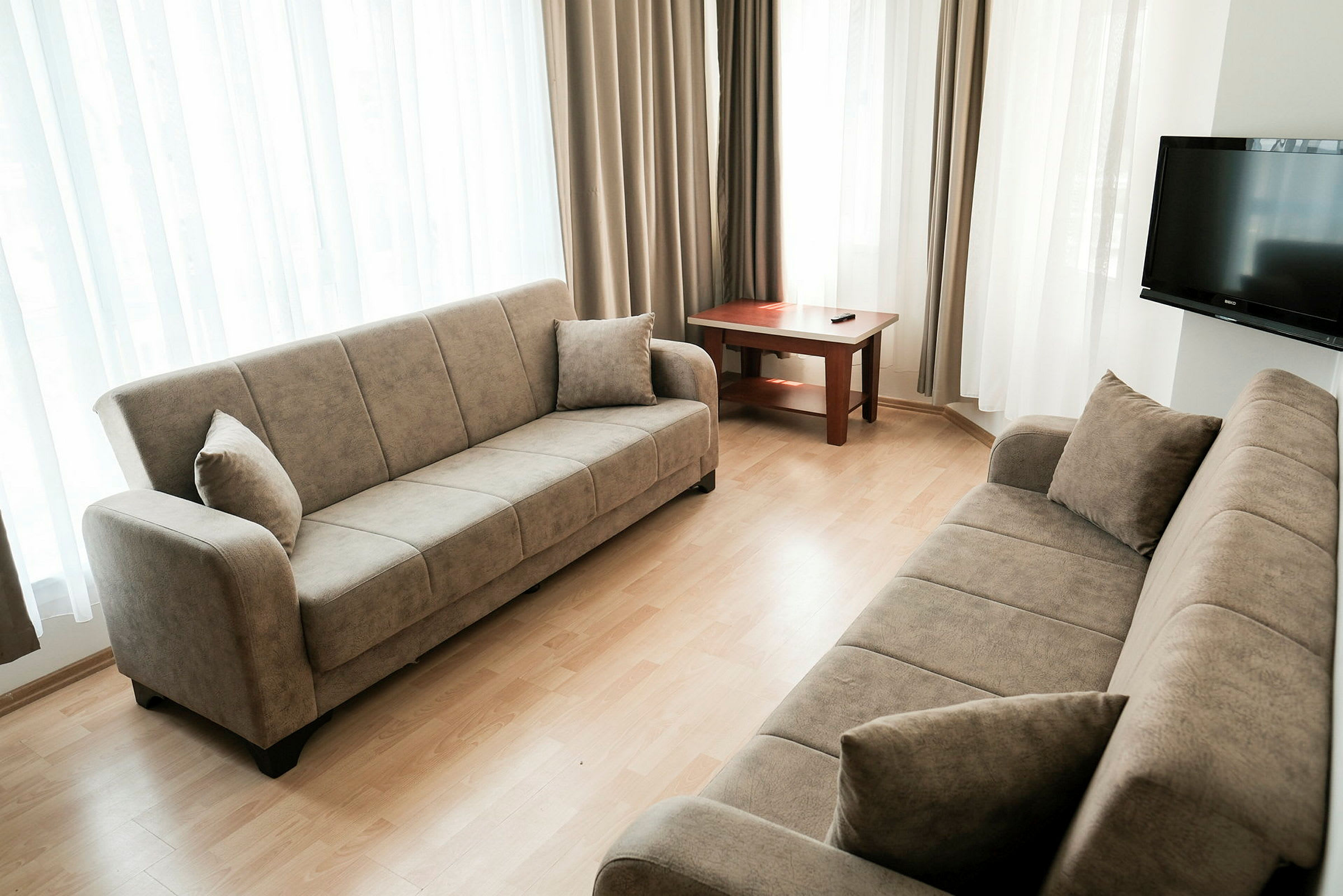 Olohuone, jossa kaksi beigeä sohvaa, puupöytä ja seinällä oleva TV. Huoneessa on vaaleat verhot ikkunoiden edessä. (Tekoälyn luoma teksti)