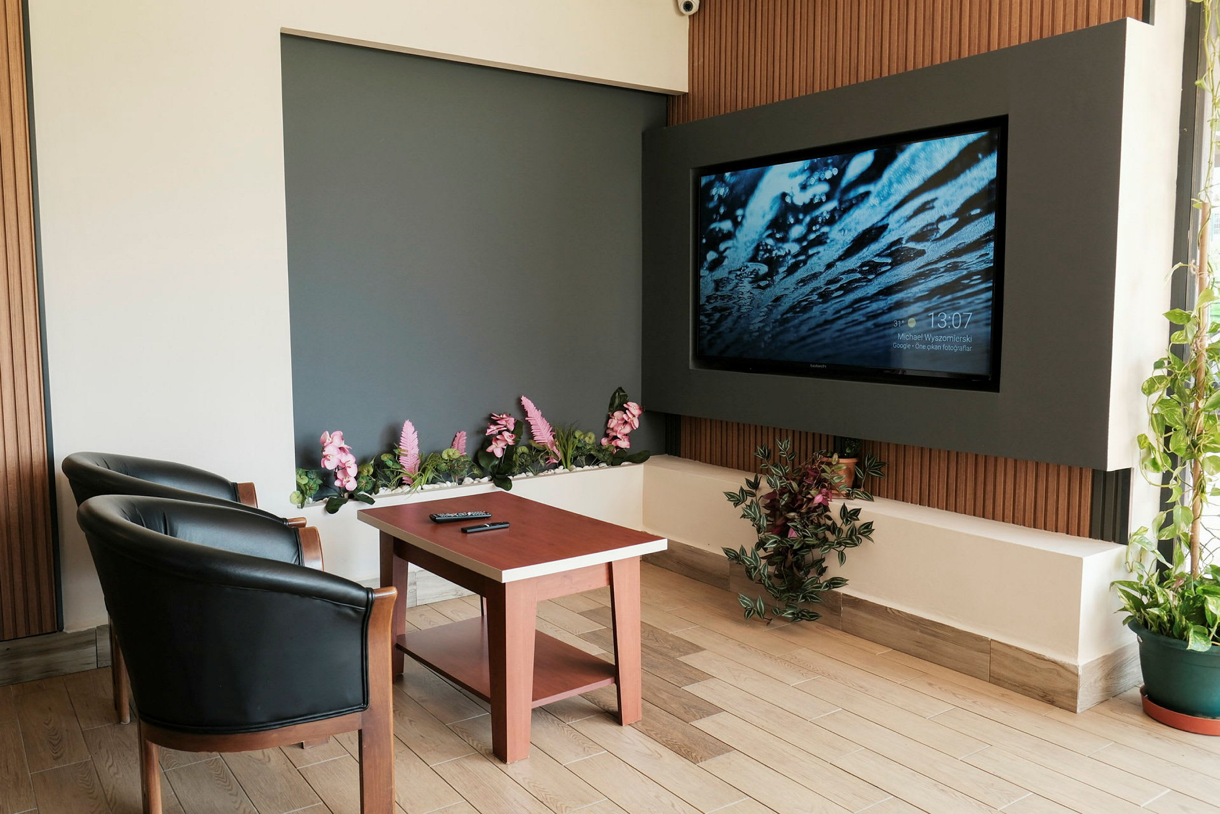 Huone, jossa on kaksi mustaa nojatuolia, puinen pöytä ja suuri seinään kiinnitetty televisio. Ympärillä on koristeellisia kukkia ja kasveja. (Tekoälyn luoma teksti)