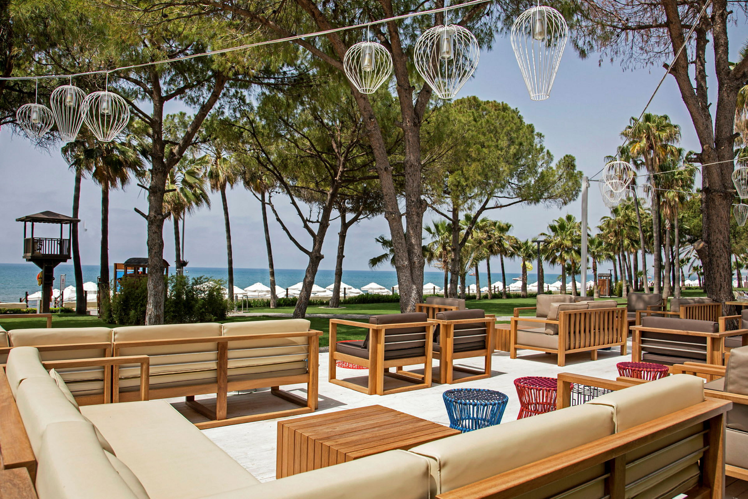 Ulkoilma istuma-alue, jossa sohvia ja pöytiä, taustalla puita, meri ja aurinkovarjoja. (Tekoälyn luoma teksti)