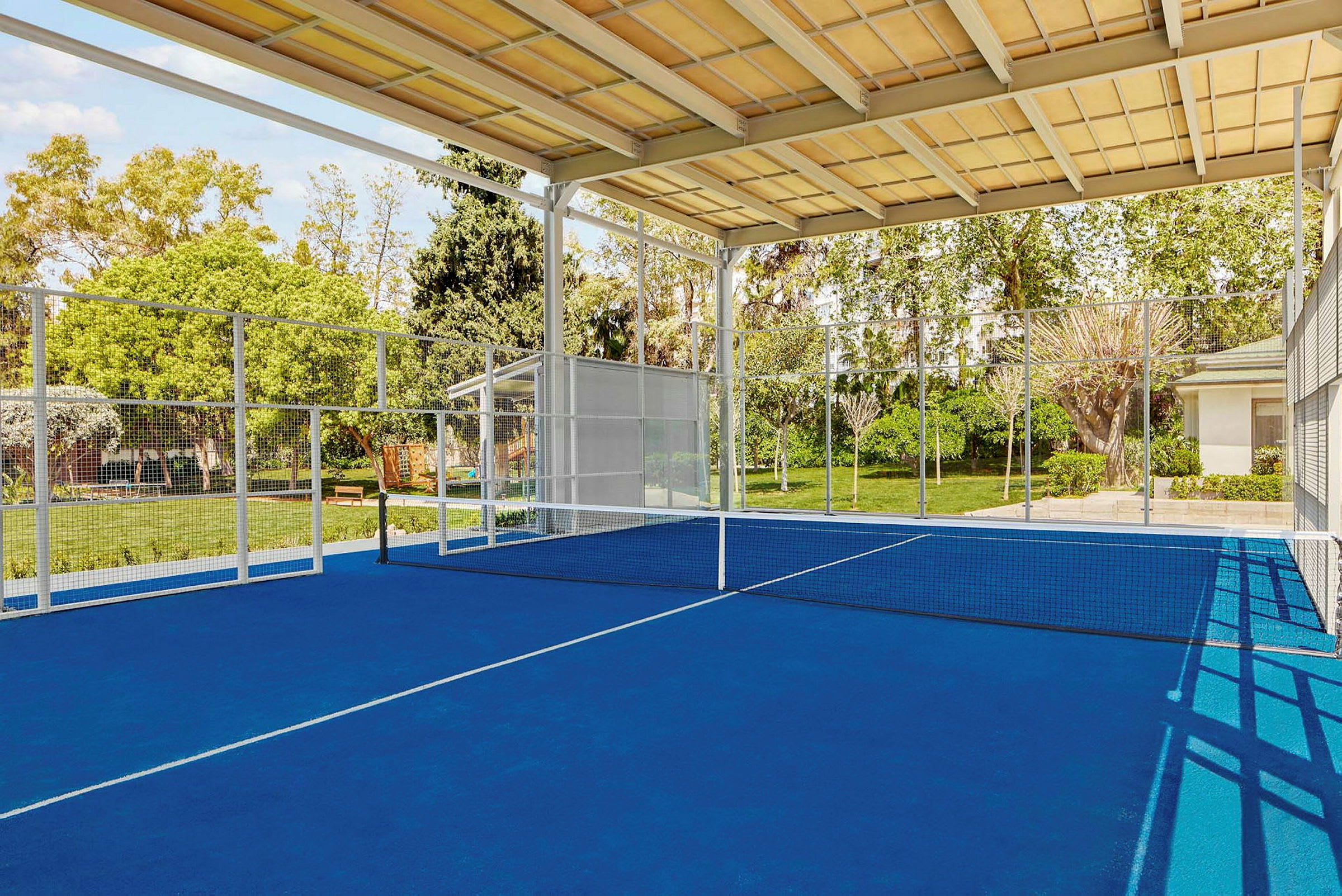 Sininen padelkenttä, jossa on verkko ja ympärillä vehreää. (Tekoälyn luoma teksti)