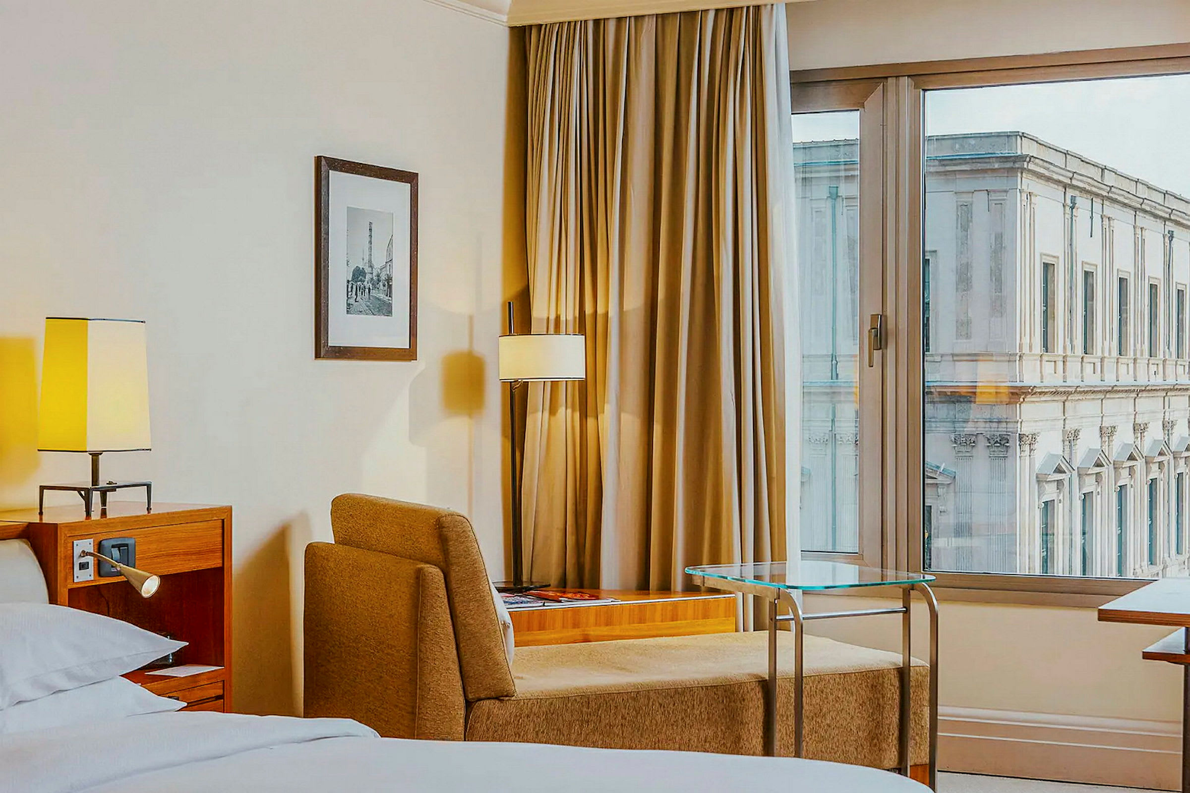 Hotellihuoneessa on sänky, nojatuoli, pöytä ja ikkuna, josta näkyy rakennus. (Tekoälyn luoma teksti)