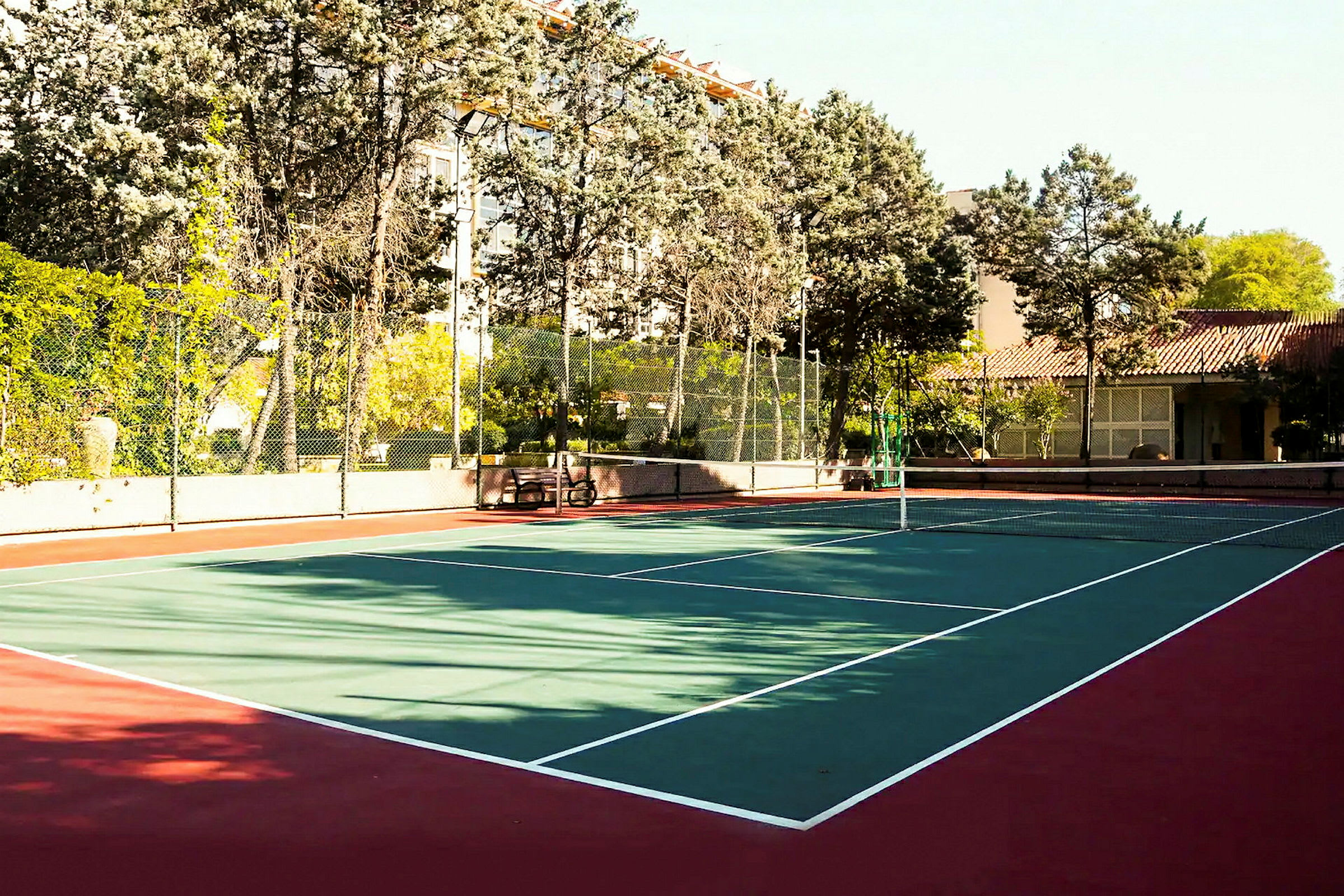 Kuva ulkokäyttöön tarkoitetusta tenniskentästä, jota ympäröivät puut ja rakennukset. (Tekoälyn luoma teksti)