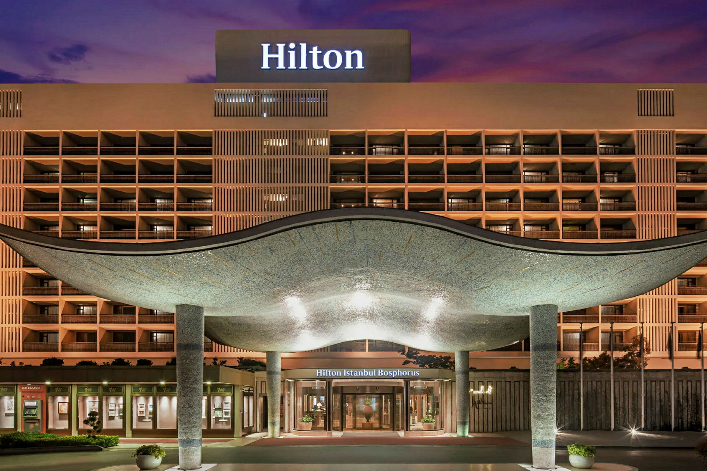 Hotellin julkisivu, jossa sisäänkäynti kaarevan katon alla, valopylväitä ja valaistu kyltti tekstillä 'Hilton'. (Tekoälyn luoma teksti)