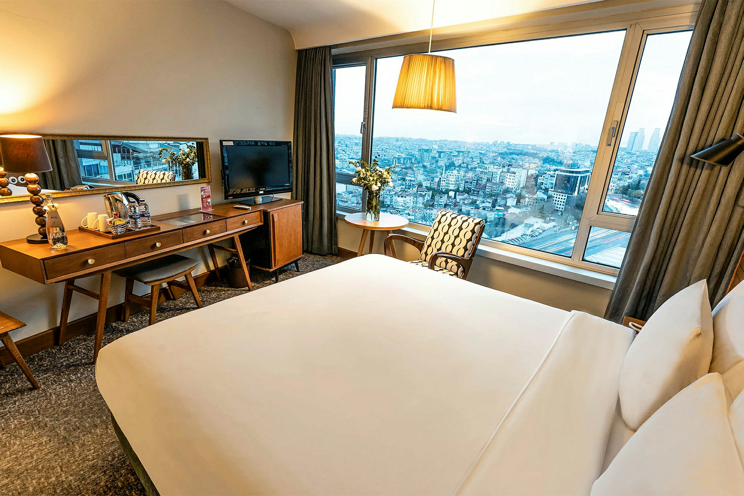 Hotellihuone, jossa on suuri sänky, kirjoituspöytä ja peili sekä iso ikkuna, josta näkyy näkymä kaupunkiin. (Tekoälyn luoma teksti)