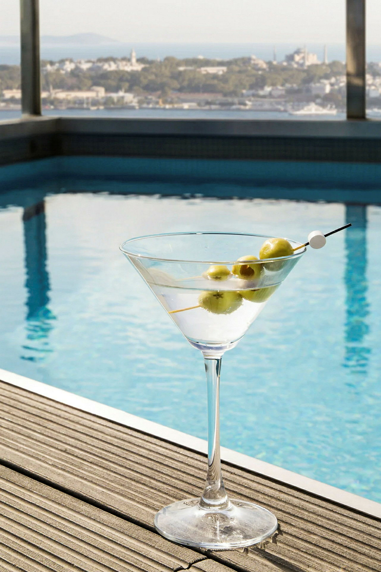 Cocktail-lasi ja oliiveja puupinnalla uima-altaan vieressä, näkymä kaupunkimaisemaan. (Tekoälyn luoma teksti)