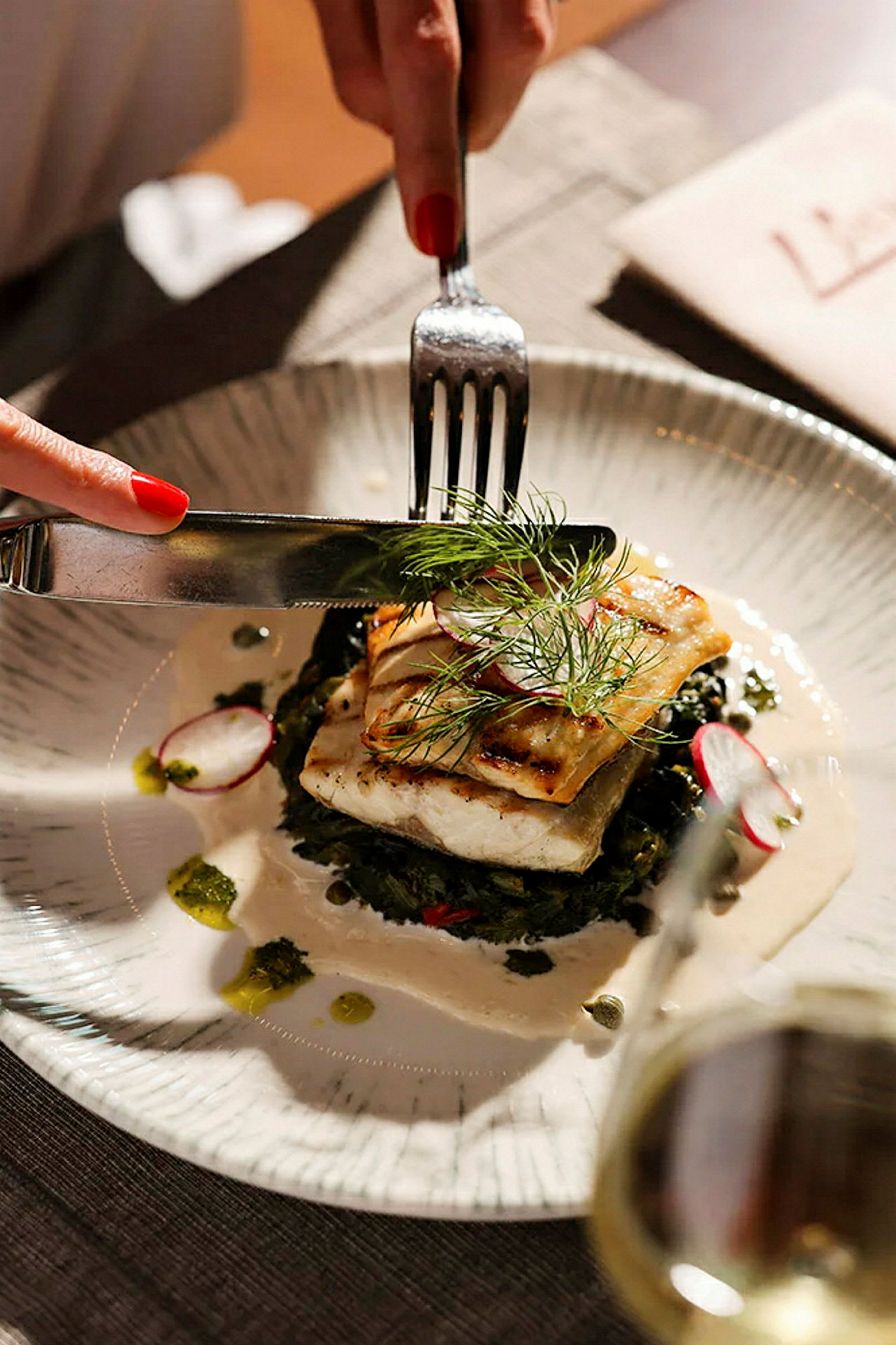 Lautanen, jolla on ateria, sisältäen grillattua kalaa, tarjoiltuna kasvisten ja kastikkeen kanssa. Ruokailuvälineitä käytetään ruoassa. (Tekoälyn luoma teksti)
