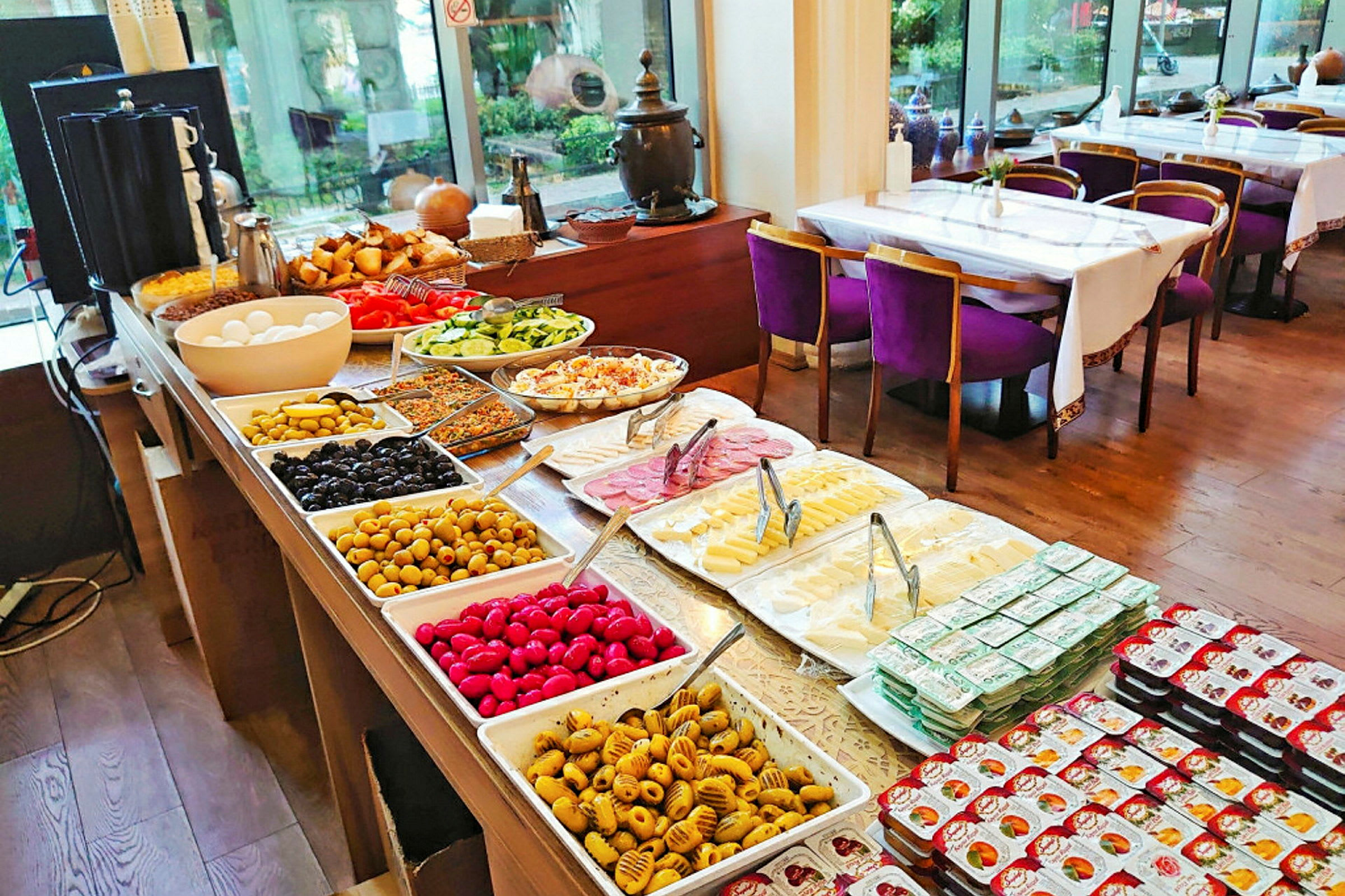 Buffet, jossa erilaisia ruokia, kulhoja ja lautasia täynnä vihanneksia, juustoja ja leikkeleitä. Taustalla ruokailutila, jossa on katettuja pöytiä ja tuoleja. (Tekoälyn luoma teksti)