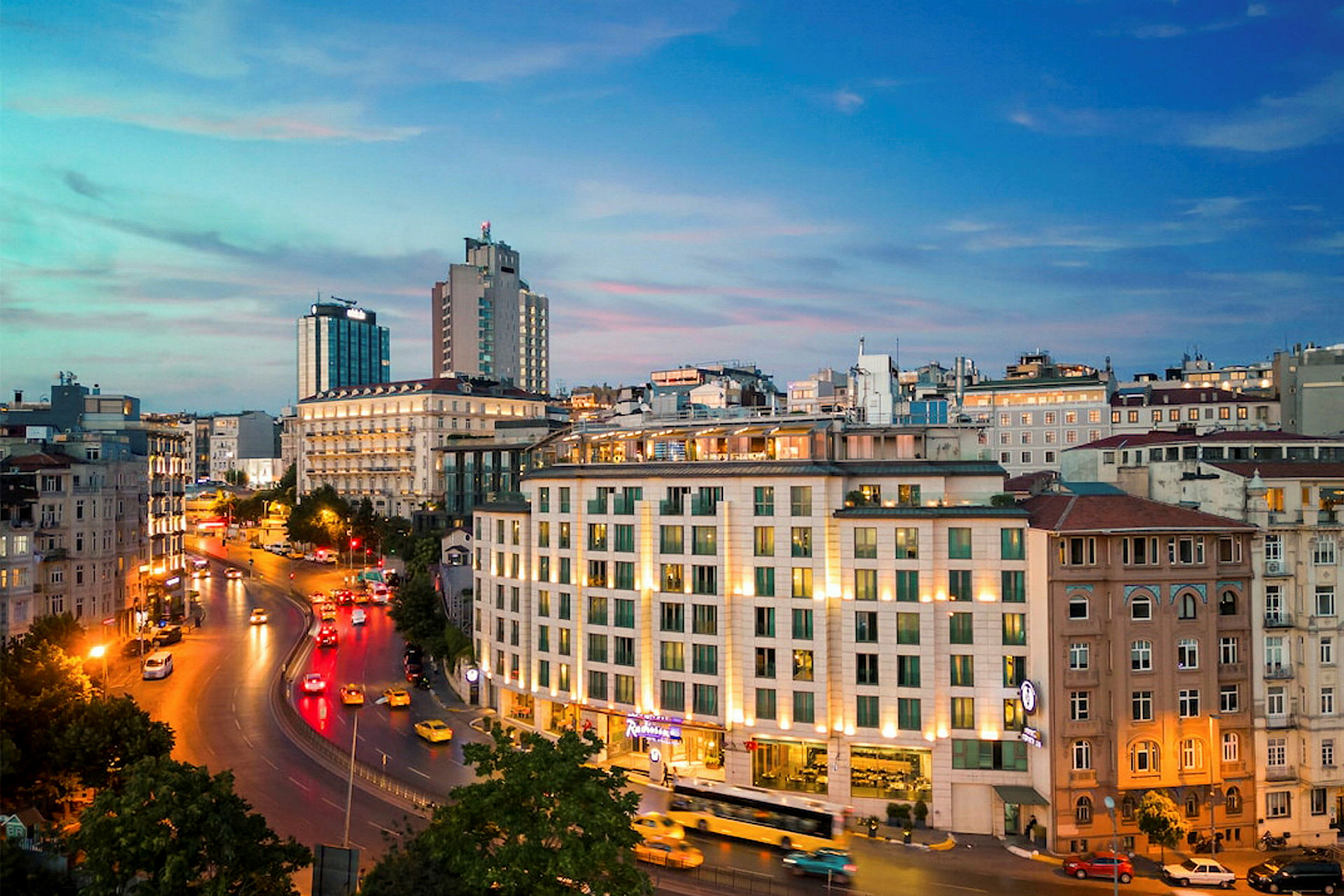 Panoraamakuva kaupunkimaisemasta hämärässä, tie, rakennuksia ja autojen valoja. (Tekoälyn luoma teksti)