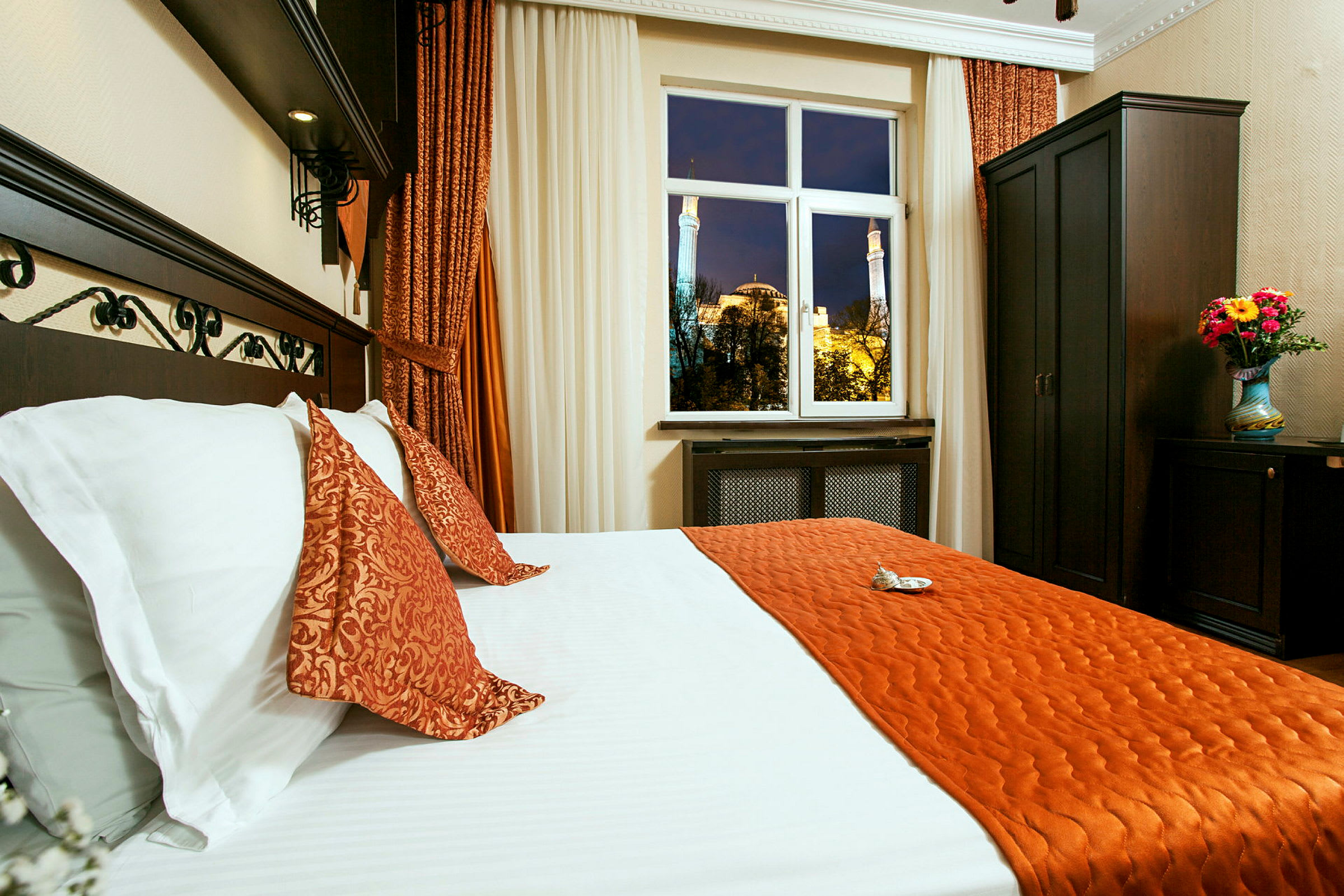 Hotellihuone, jossa sänky on valkoisilla ja oransseilla vuodevaatteilla. Ikkunasta näkyy Hagia Sofia iltanäkymä ulkoa. Tummaa kalustusta ja kukkavaasi näkyvät huoneessa. (Tekoälyn luoma teksti)